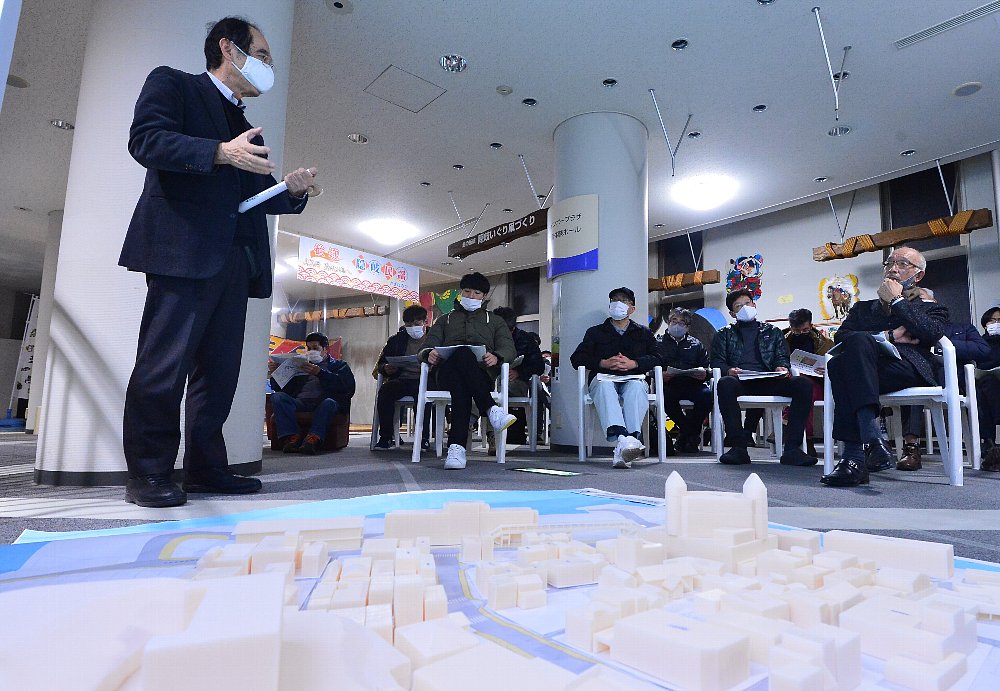 模型をもとに話をしている男性の話を椅子に座って聞いている人たちの写真