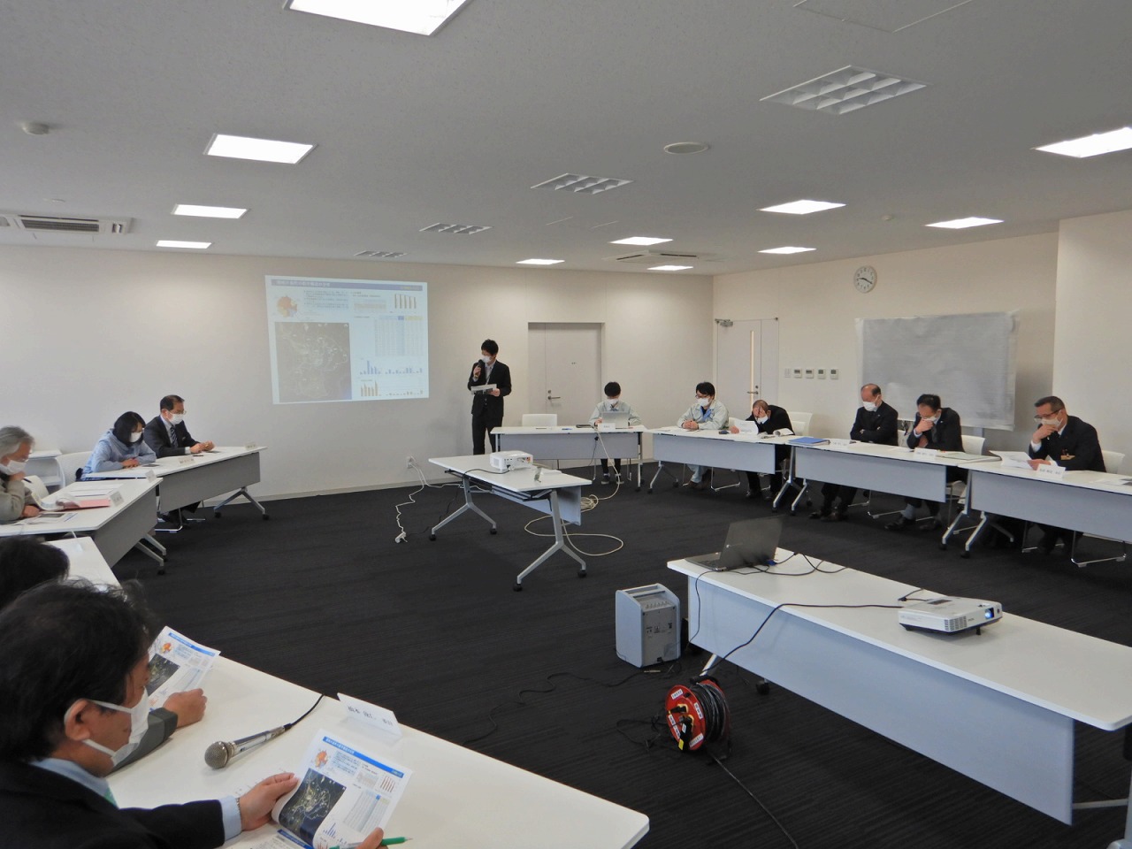 プロジェクターで映した画面をもとに話す男性と資料を見ている人たちの会議中の写真
