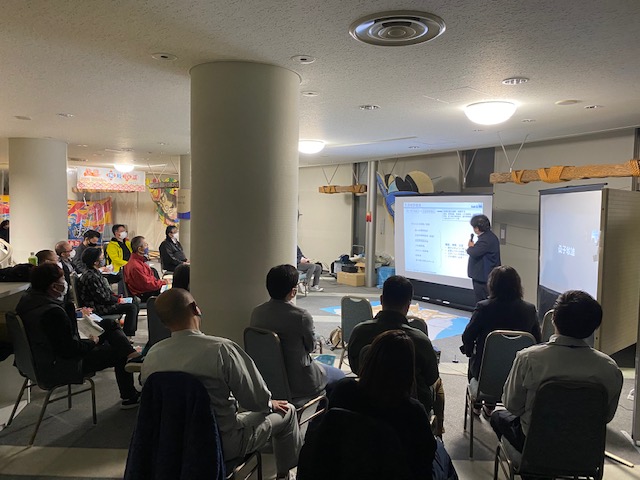 スクリーンをもとに話している人の話を座って聞いている人たちの写真