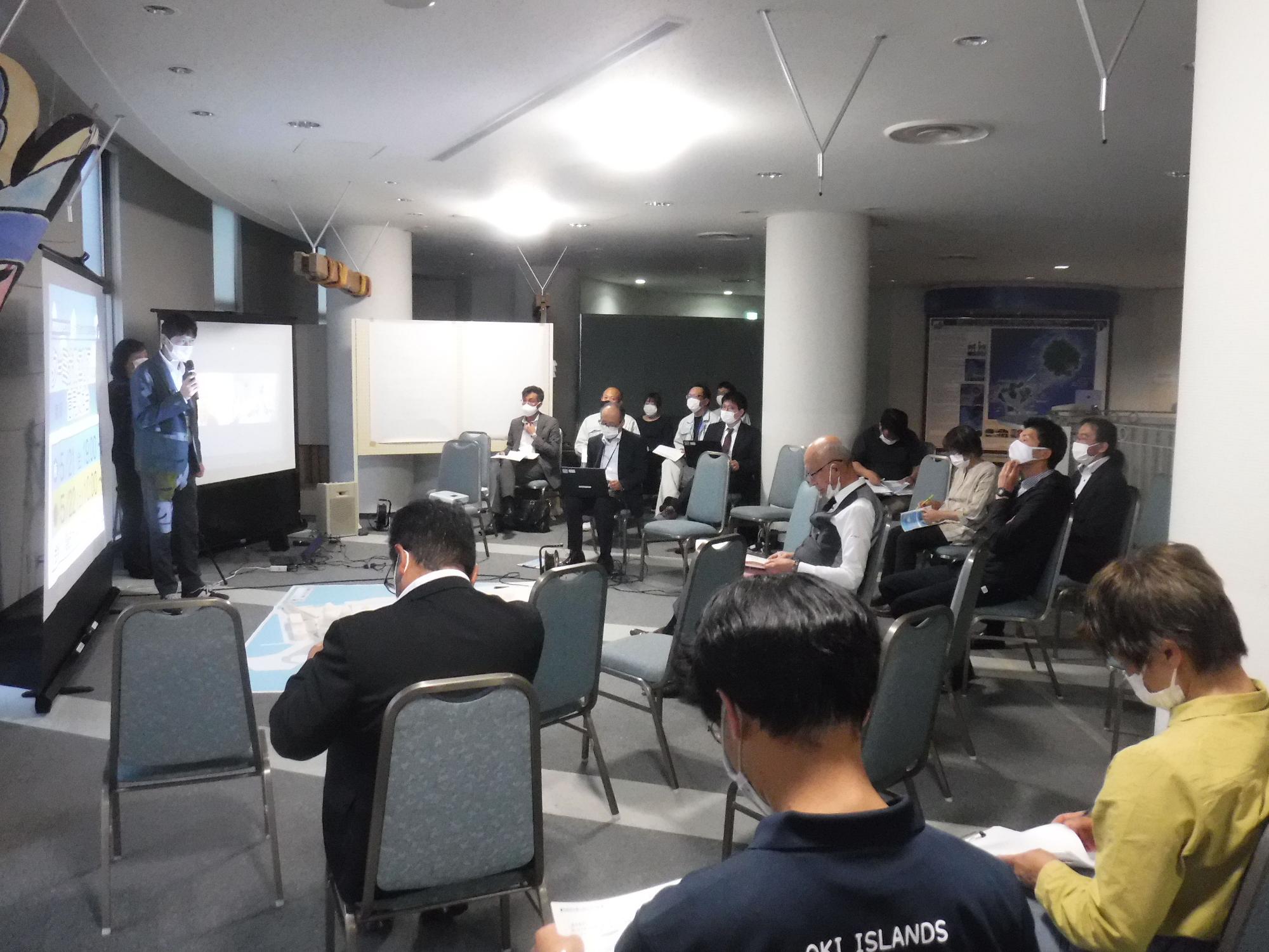 スクリーンの前で話をしている人と椅子に座って資料を見ている人たちの写真