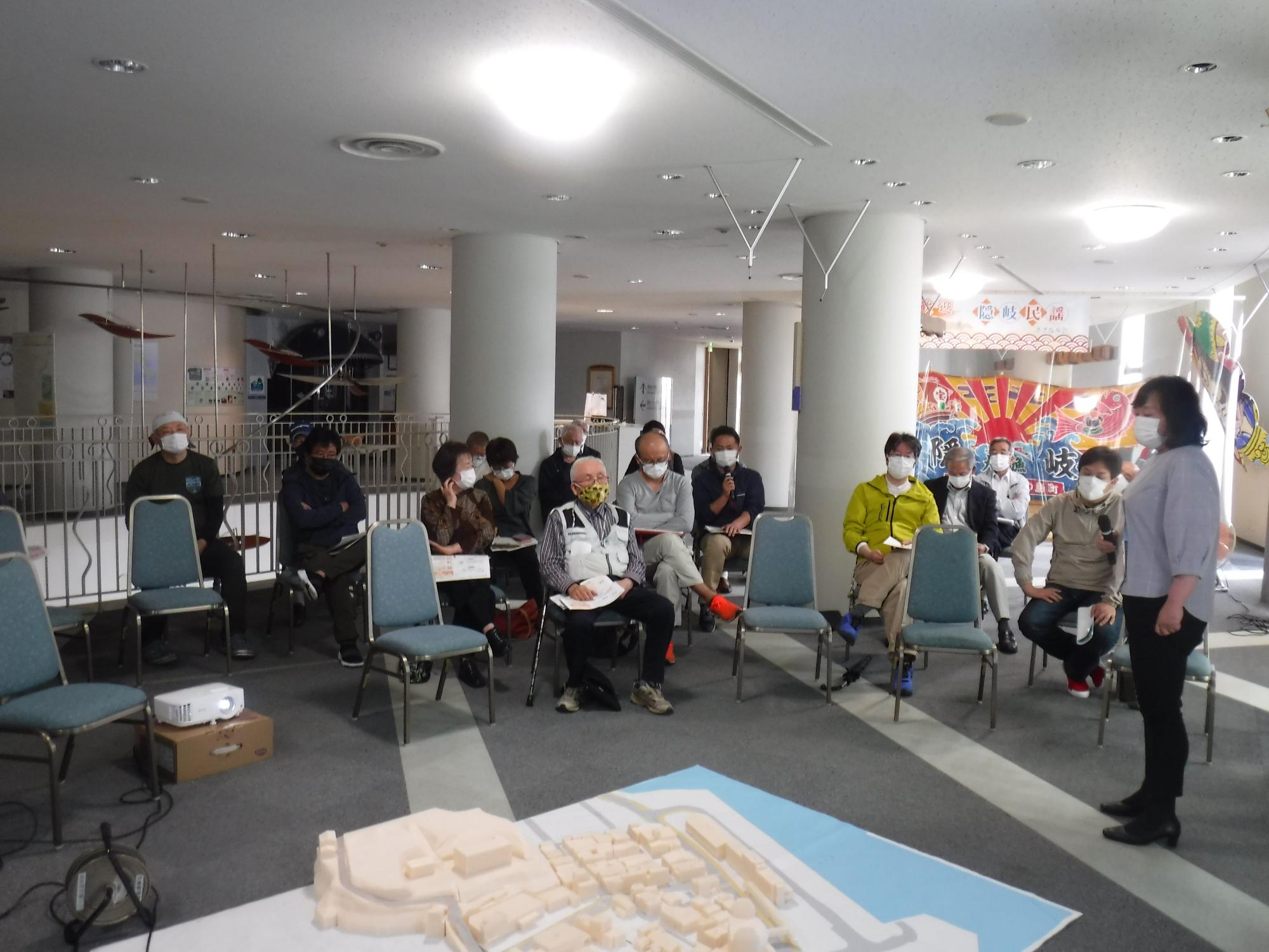 模型を見ながら話す女性を椅子に座って見ている人たちの写真