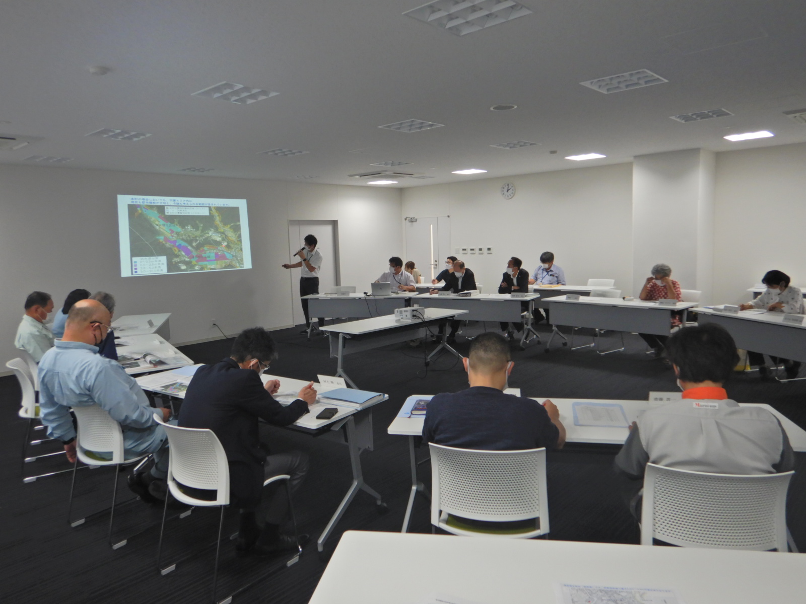 プロジェクターで映した地図を見ながら話す男性と席で資料を見ている人たちの写真