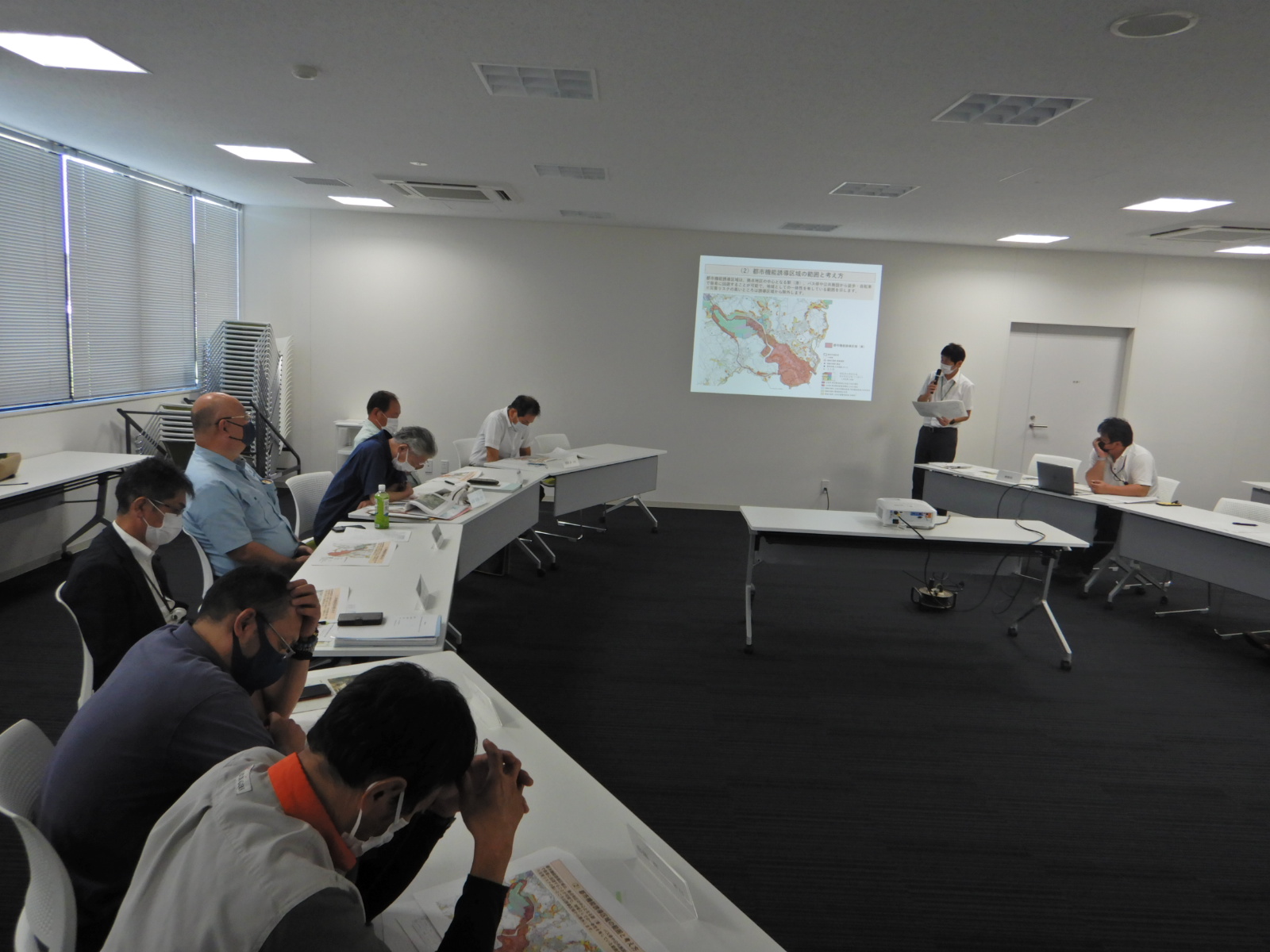 プロジェクターで映した画面を見て話す人と机の資料を見て頭をかかえる人たちの写真