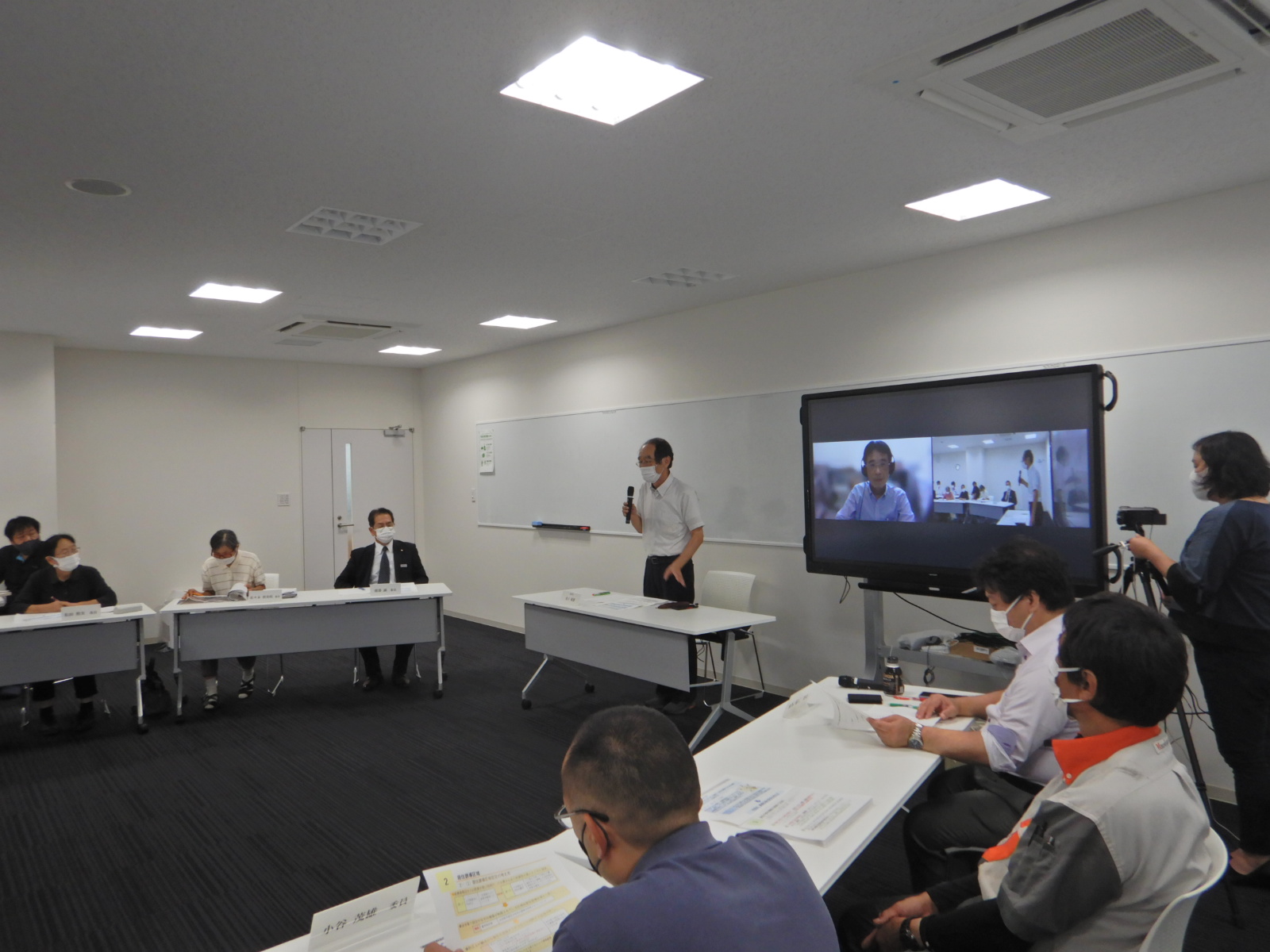 モニターの横で話す男性と席で資料を見ている人たちの写真