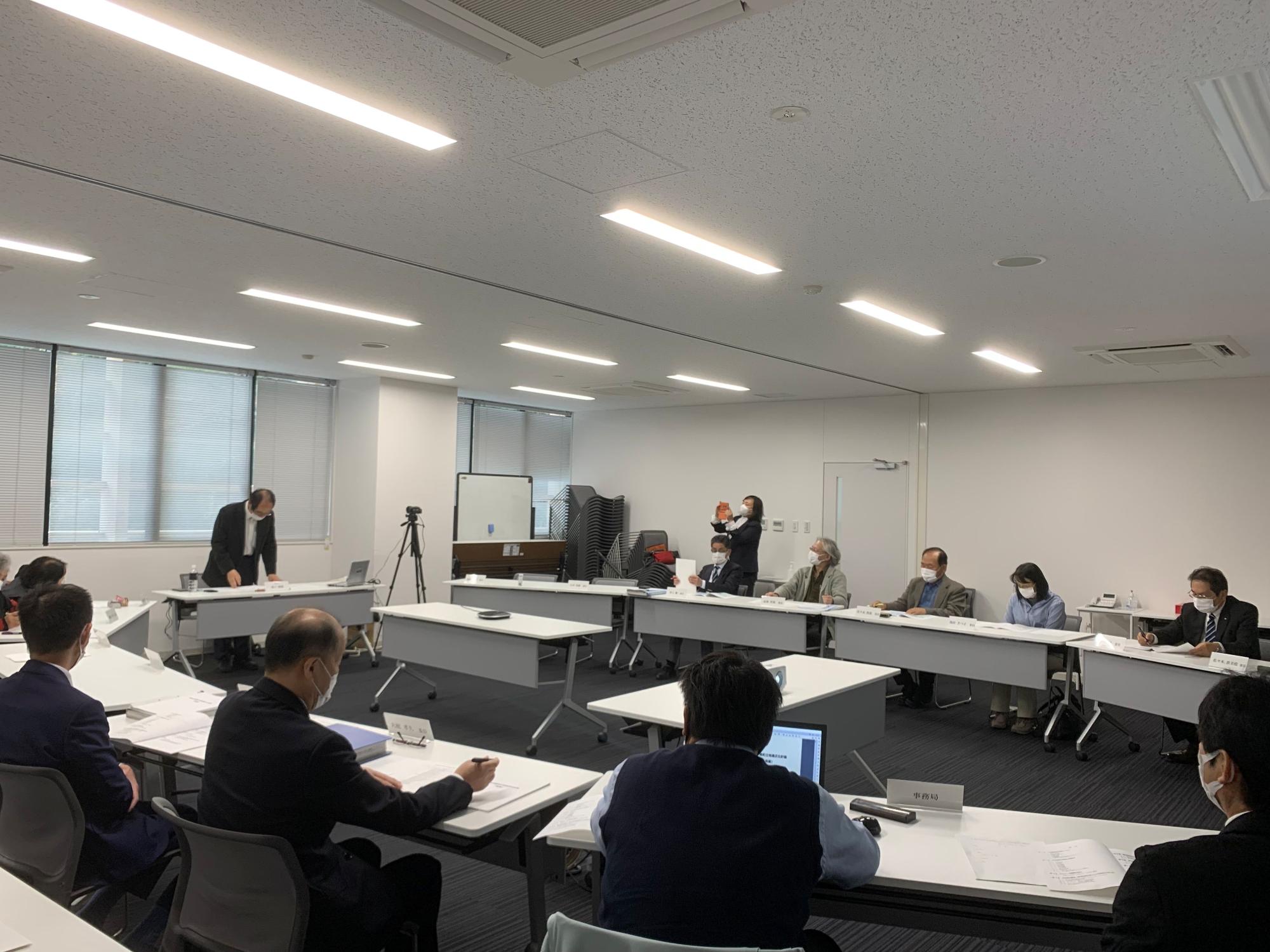 立って資料を見ている男性を座って見ている人たちの写真