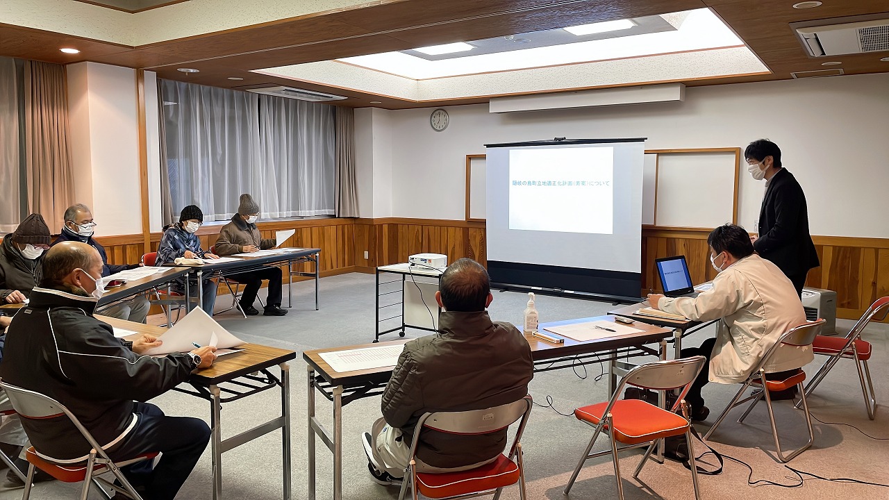 前方にスクリーンがある室内でテーブルに座った複数の人たちが手元の資料を見ている写真