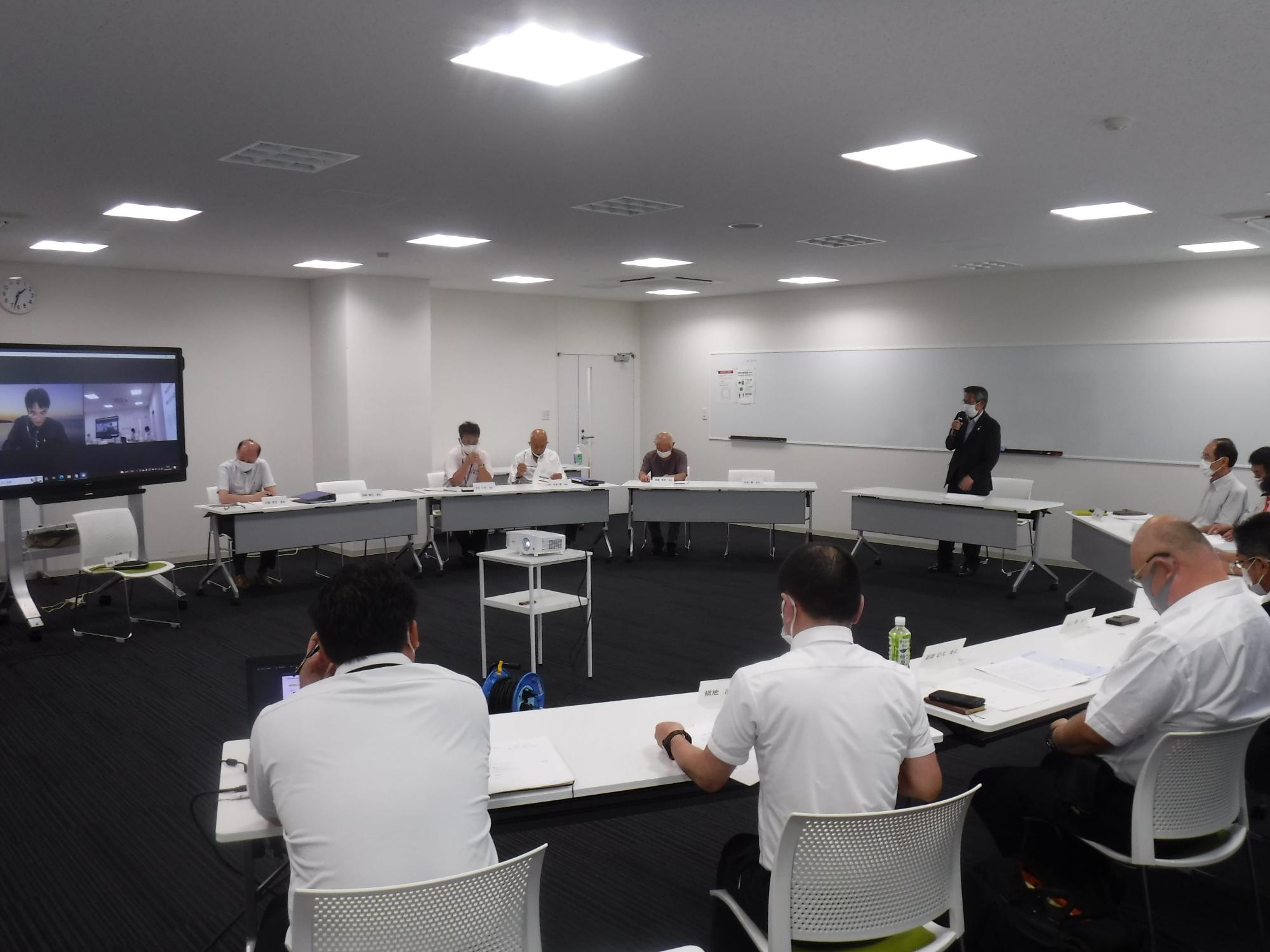 デスクの前で立つスーツ姿の男性の話を円形に並んだ席に座って聞く人たちの写真