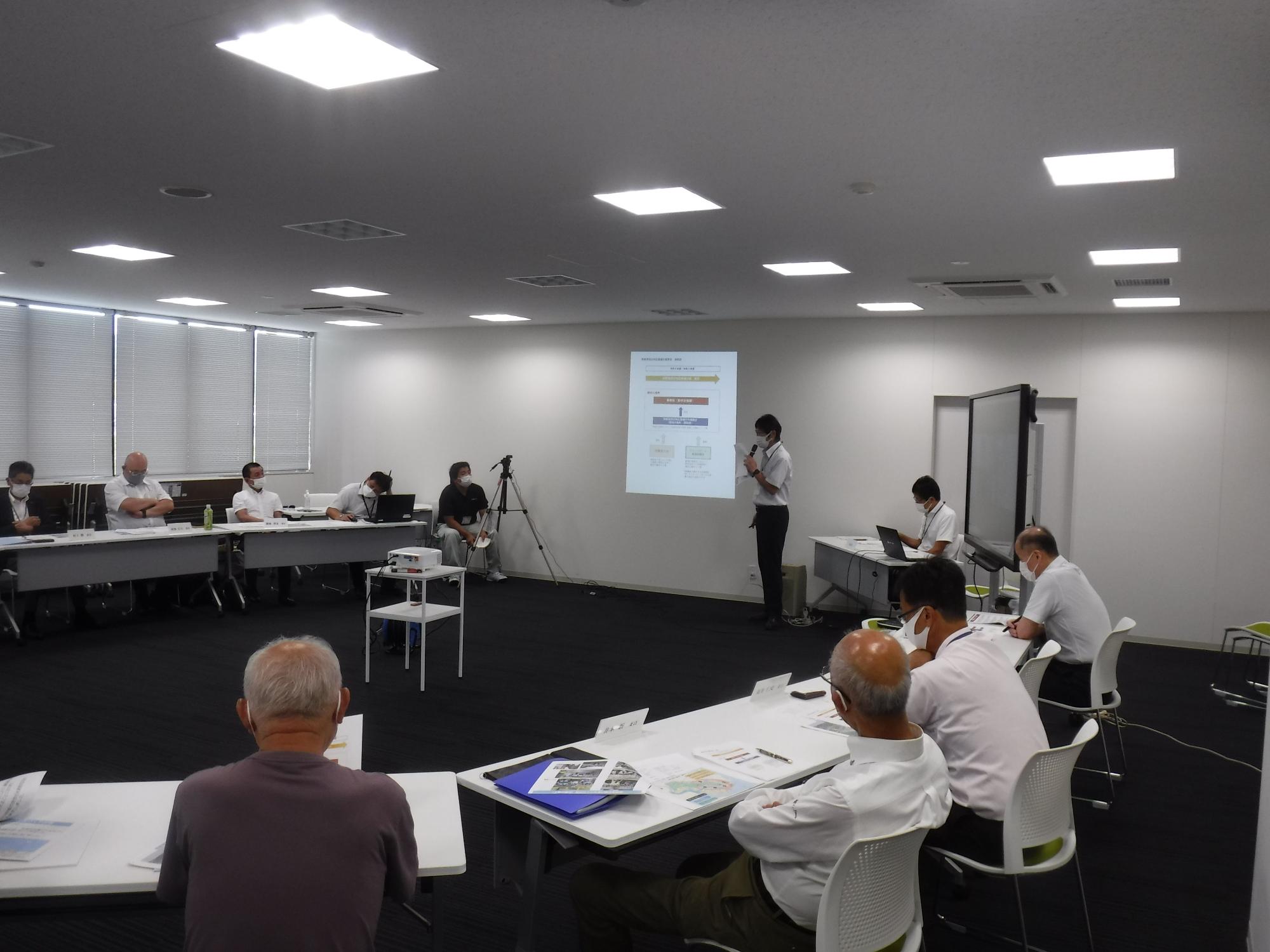 マイクを手に持ち立って話す男性を円形に並んだ席から見ている人たちの写真