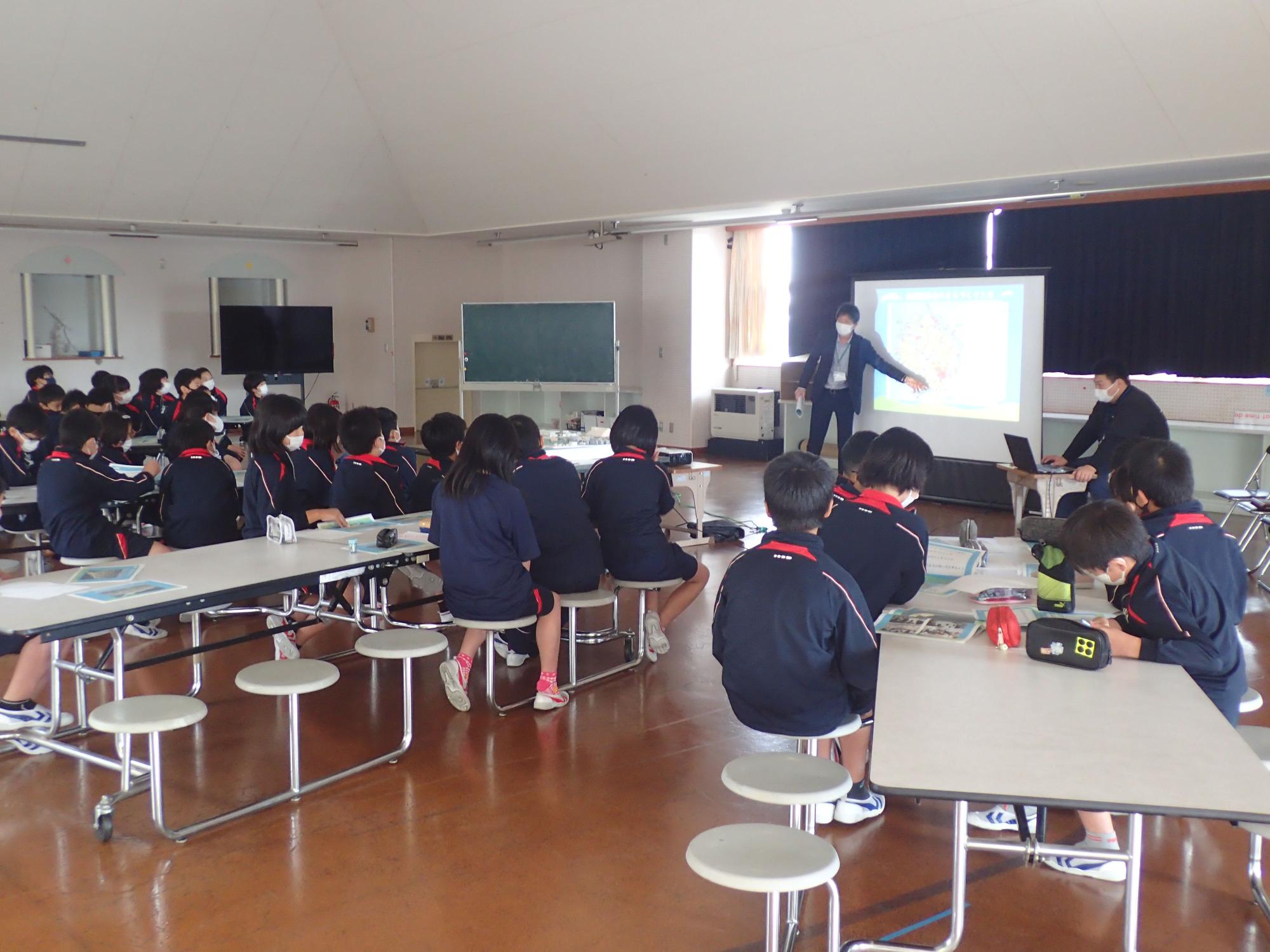 スクリーンを用いて話す先生を見る生徒の後ろ姿の写真