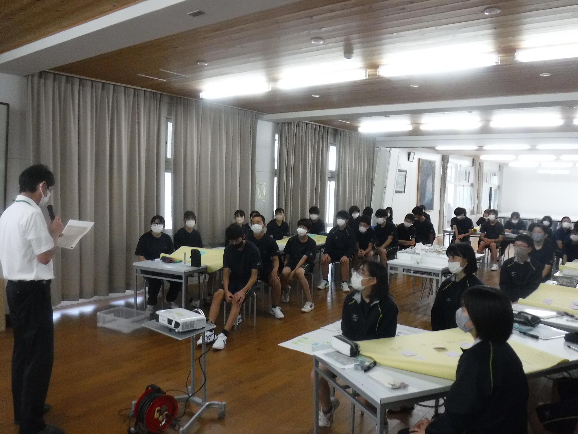 前に立って話す男性を見ているマスク姿の生徒たちの写真