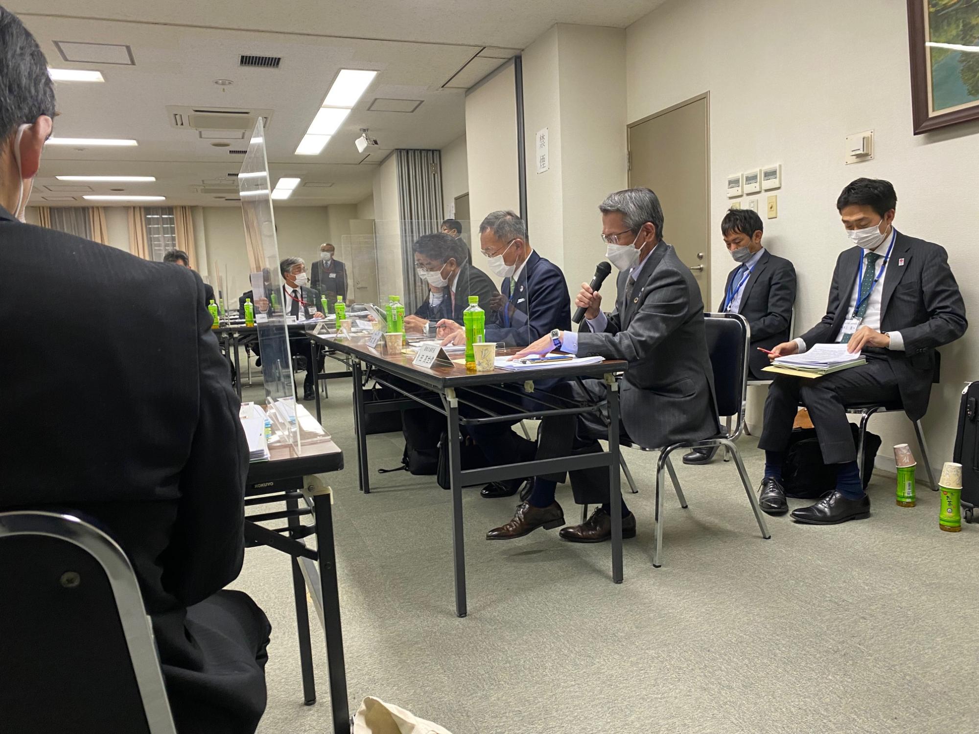 机の資料を見ながら話すスーツ姿の男性とその後ろで膝に置いた資料を確認しているスーツ姿の男性の写真