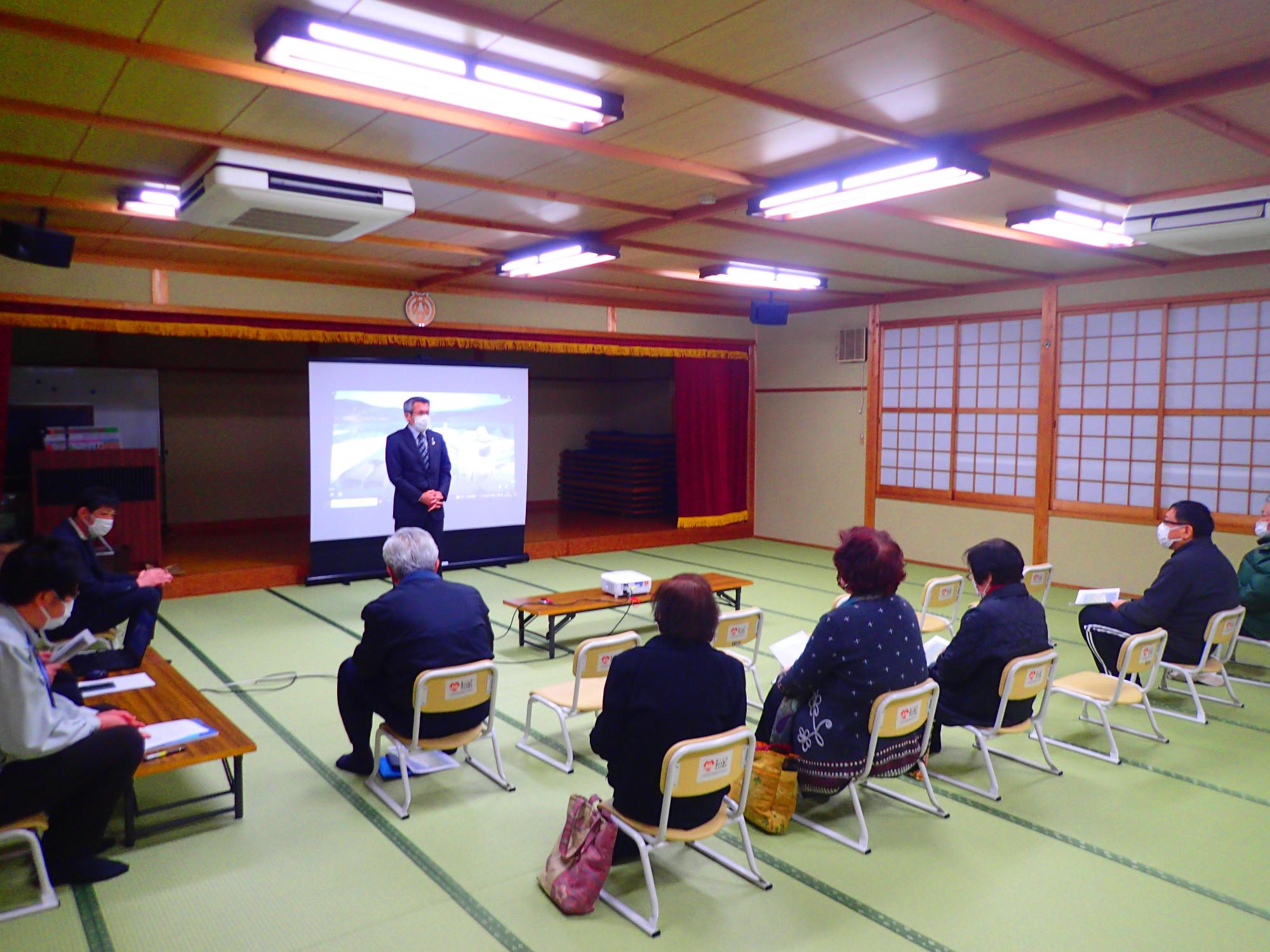 座敷の室内で椅子に座った人たちの前で男性が立っている写真