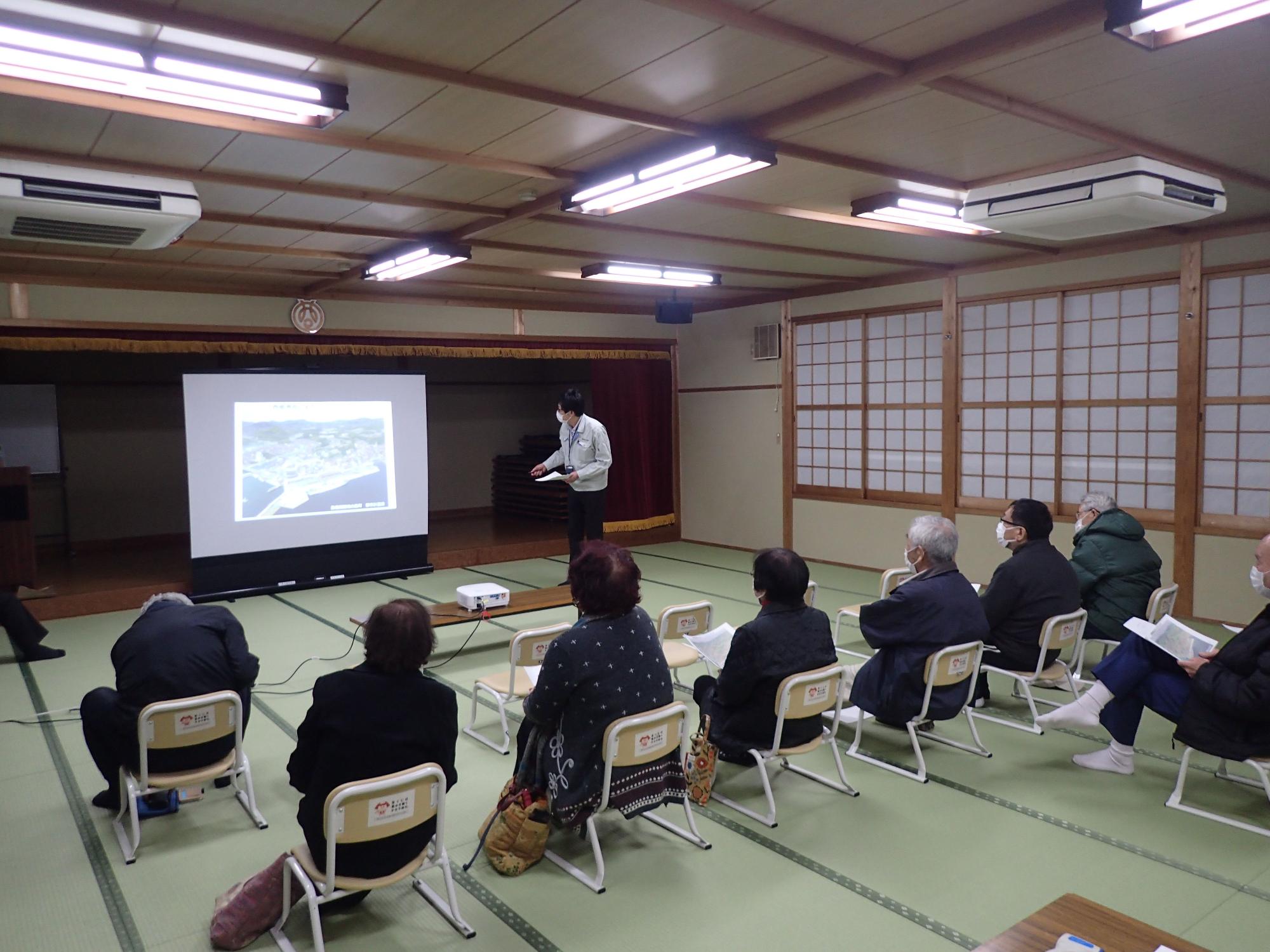 座敷の室内で椅子に座った人たちが前方のスクリーンを見ている写真