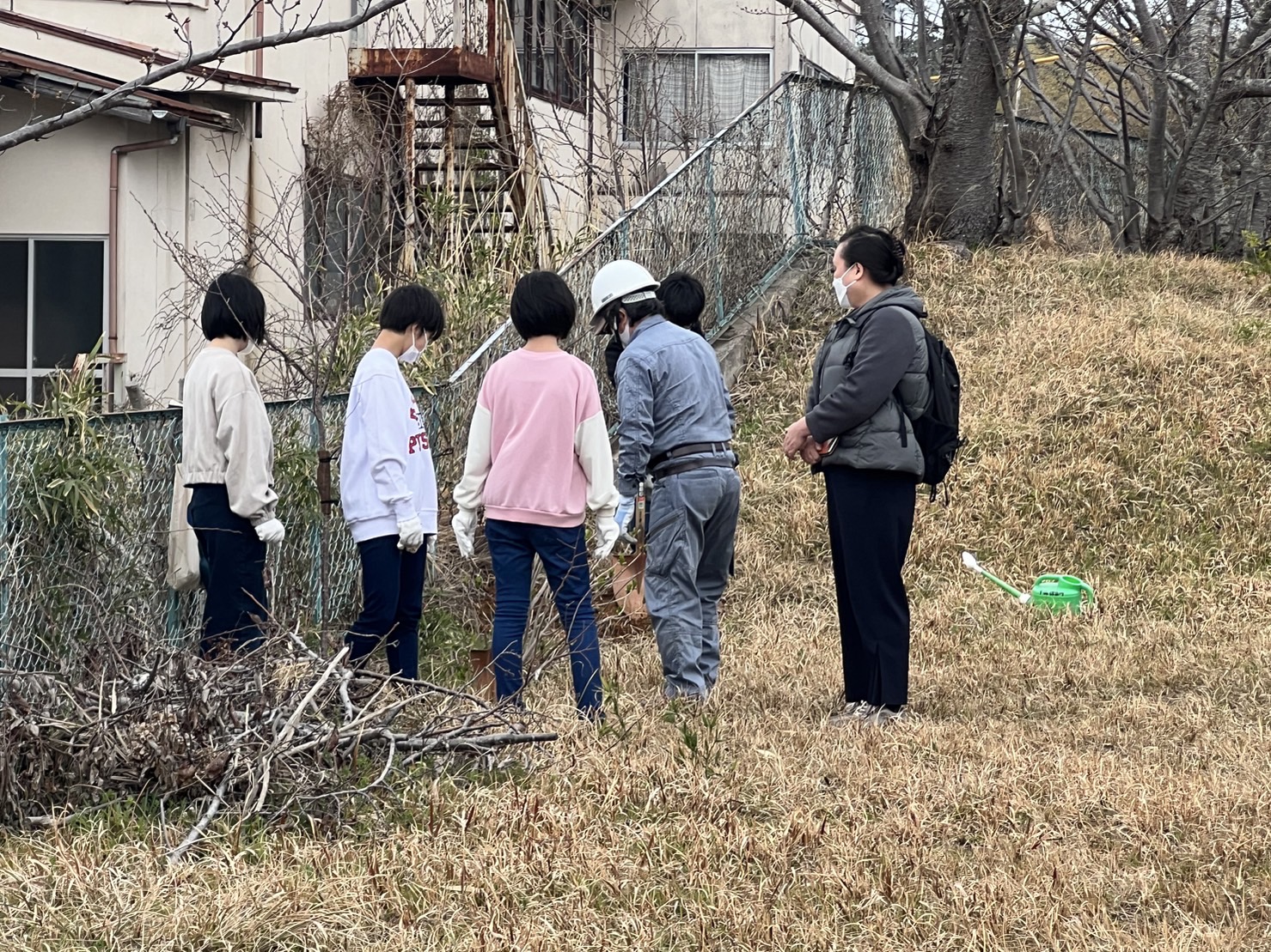 鉄柵がある公園の端でシャベルを手に作業する男性と男子生徒を見守る児童達と生徒達の写真