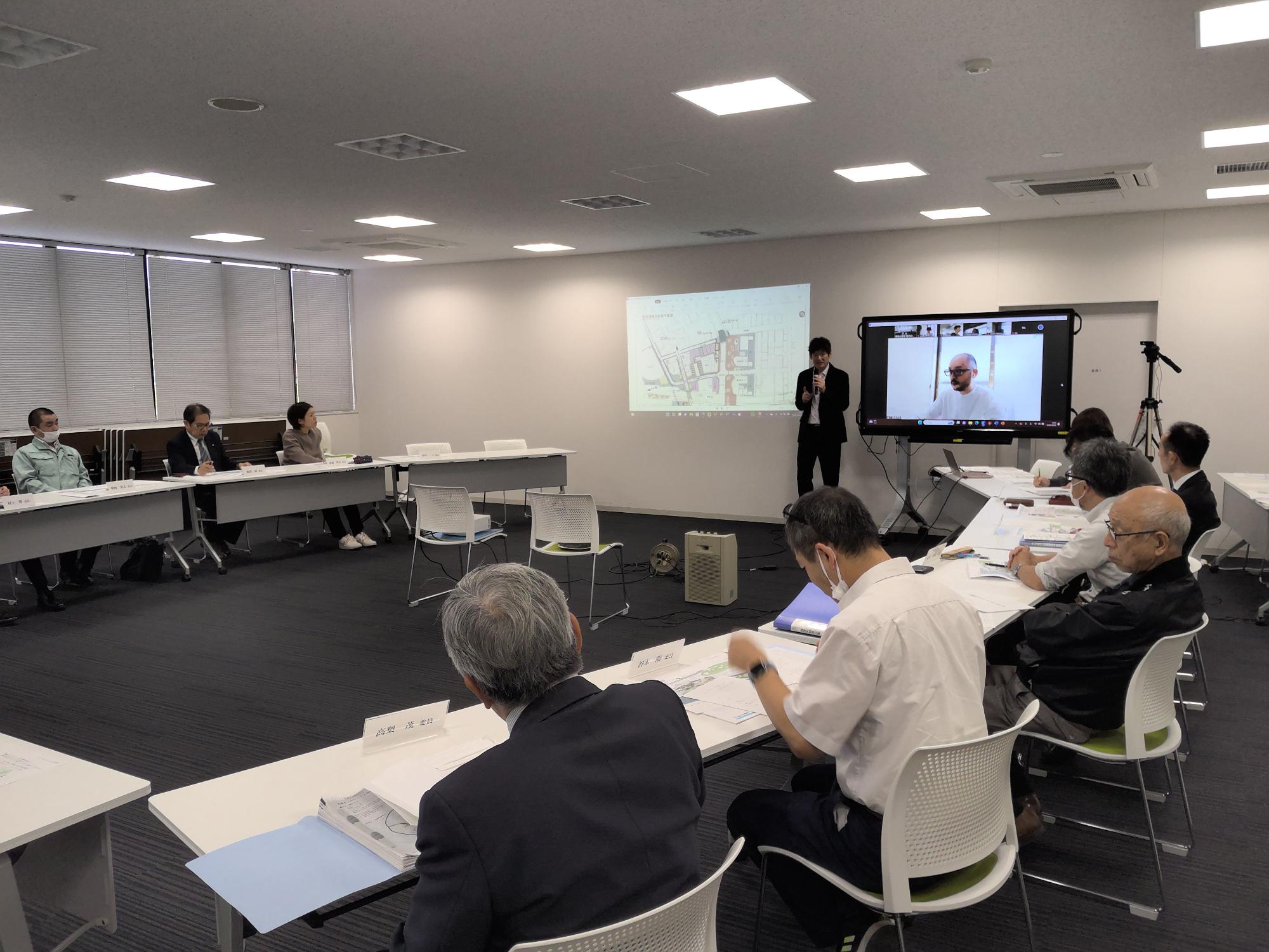 モニターの横に立つ男性の話を資料を見ながら聞いている人たちの写真