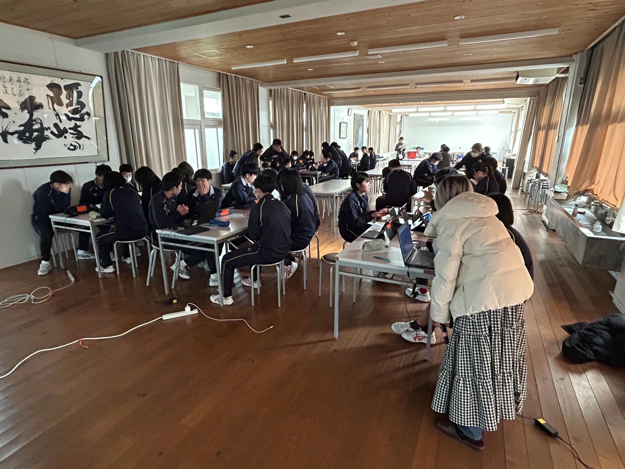 ジャージ姿の生徒たちが広い教室の席に座っている写真