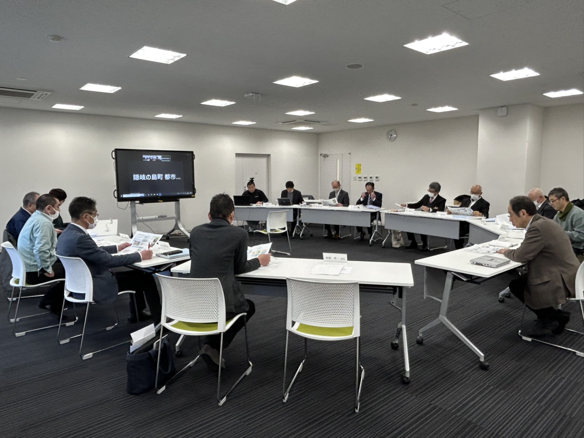 モニターを前に円形に並んだ席に座って会議する人々の写真