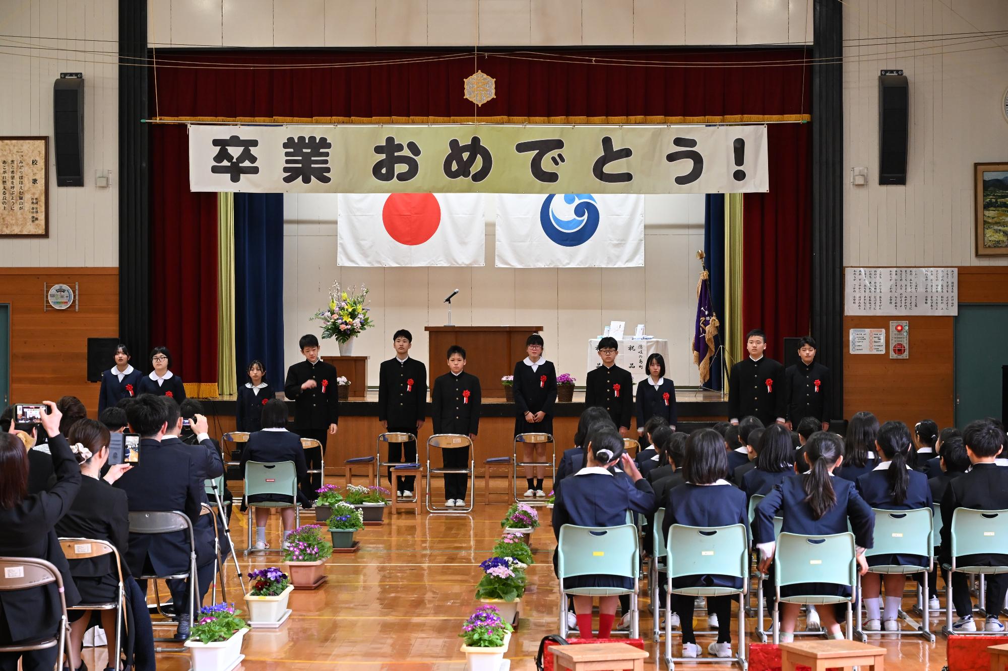 6年生が卒業式にて起立し、校歌を斉唱しているのを保護者と在校生が見ている様子が映った写真