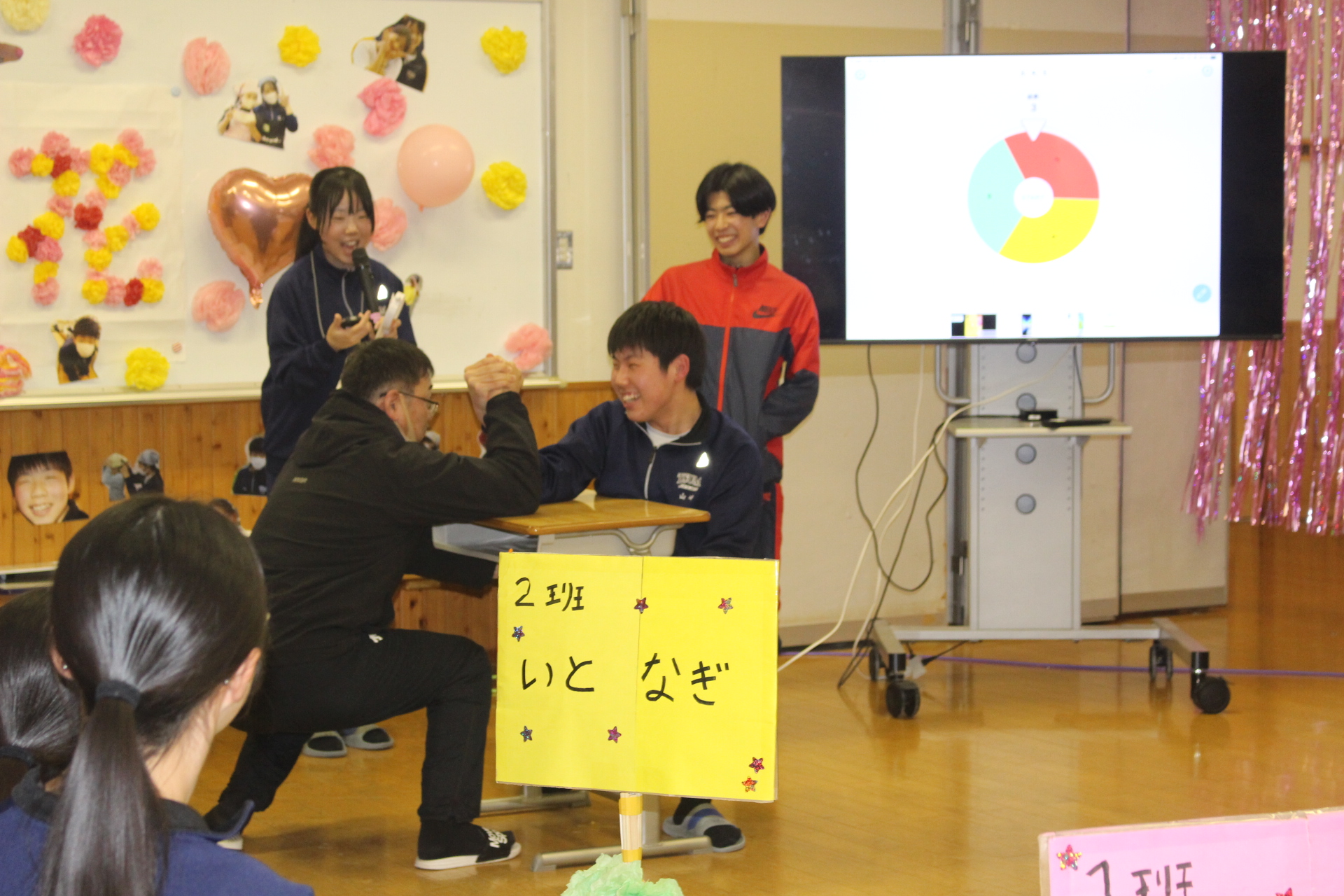 3年生を送る会で腕相撲をする男子生徒2名とそれを見ている児童たち