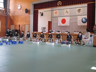 「祝入学」と幕がかけられた体育館のステージの前で、マスクを付けた制服姿の新入生が横一列に座っている写真