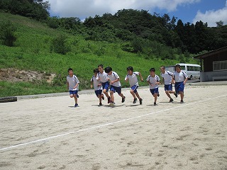 校庭のトラックの上を、体操着姿の児童たちが一列になって走っている様子の写真