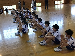 体育館でマスクを付けた児童が横二列に体育座りで座っている様子の写真