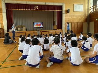 体育館で正面にモニターを使って資料を使いながら話す講師の方の話を、座って熱心に聞いている児童たちの写真