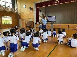 体育館の正面に講師の方が立ち、手を上げながら座っている児童たちに説明している様子の写真