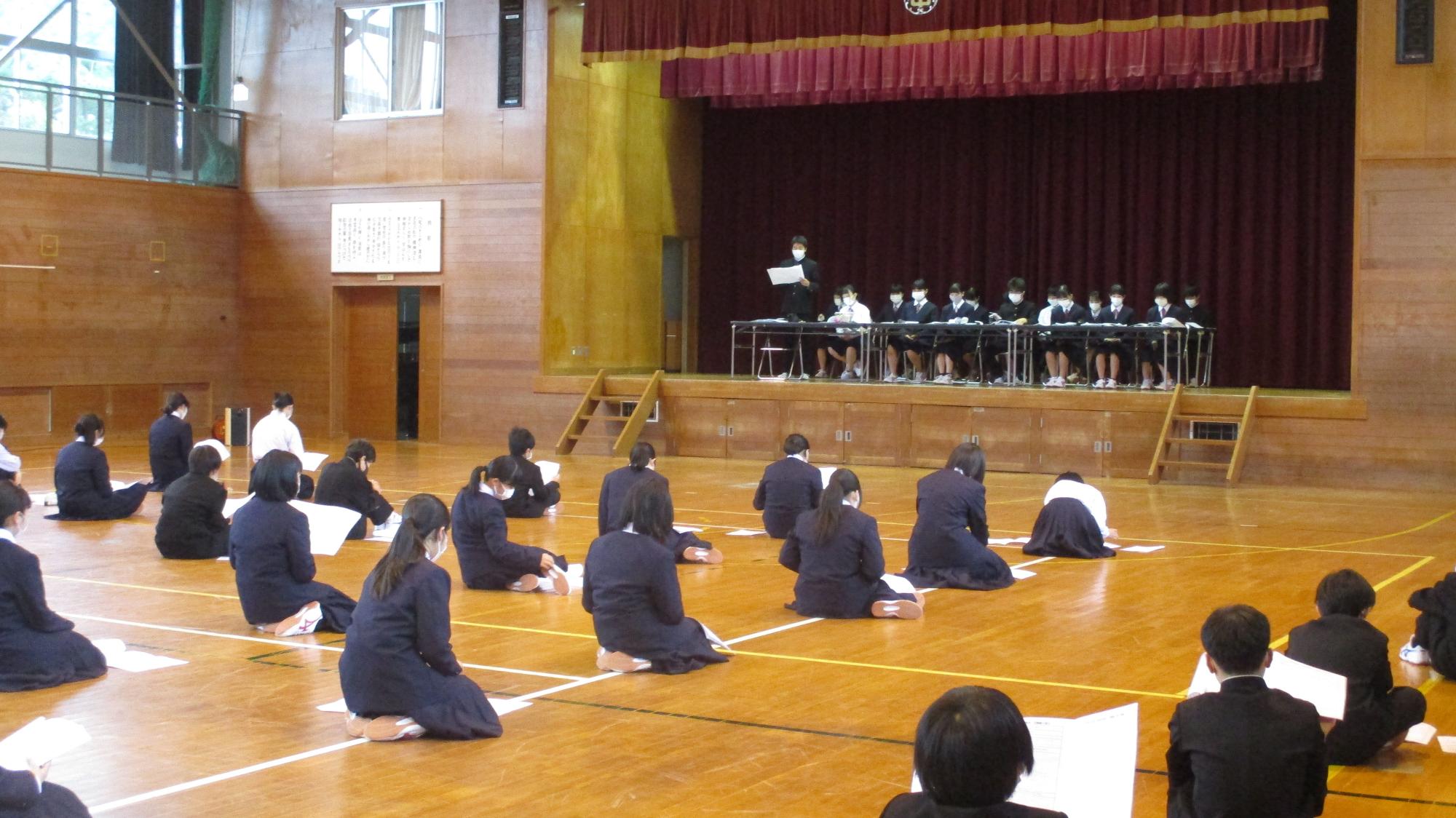 体育館で生徒が互いにソーシャルディスタンスを取りながら座り、生徒総会に参加している写真