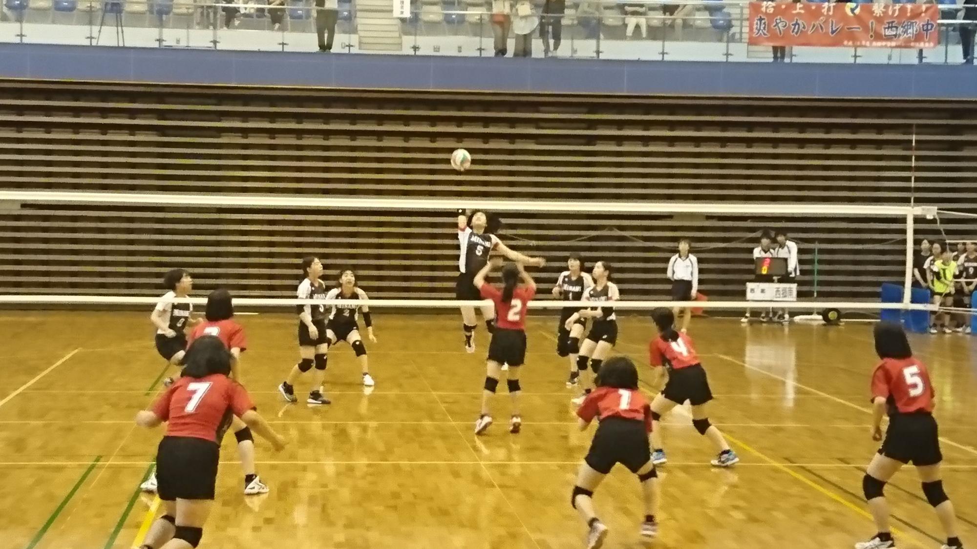 体育館のバレーコートで赤いユニフォームと白いユニフォームのチームに分かれ、白いチームの一人がスパイクを決めようとしている写真