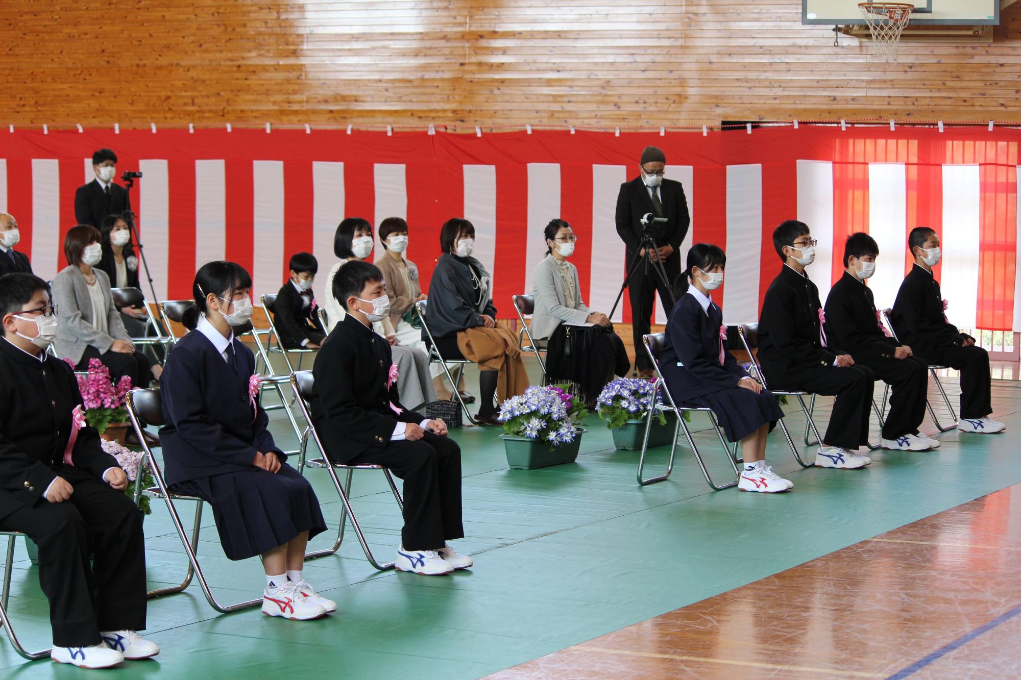 壁に紅白幕が掛けられた体育館で新中学生の男女がパイプ椅子に緊張した面持ちで座っている写真