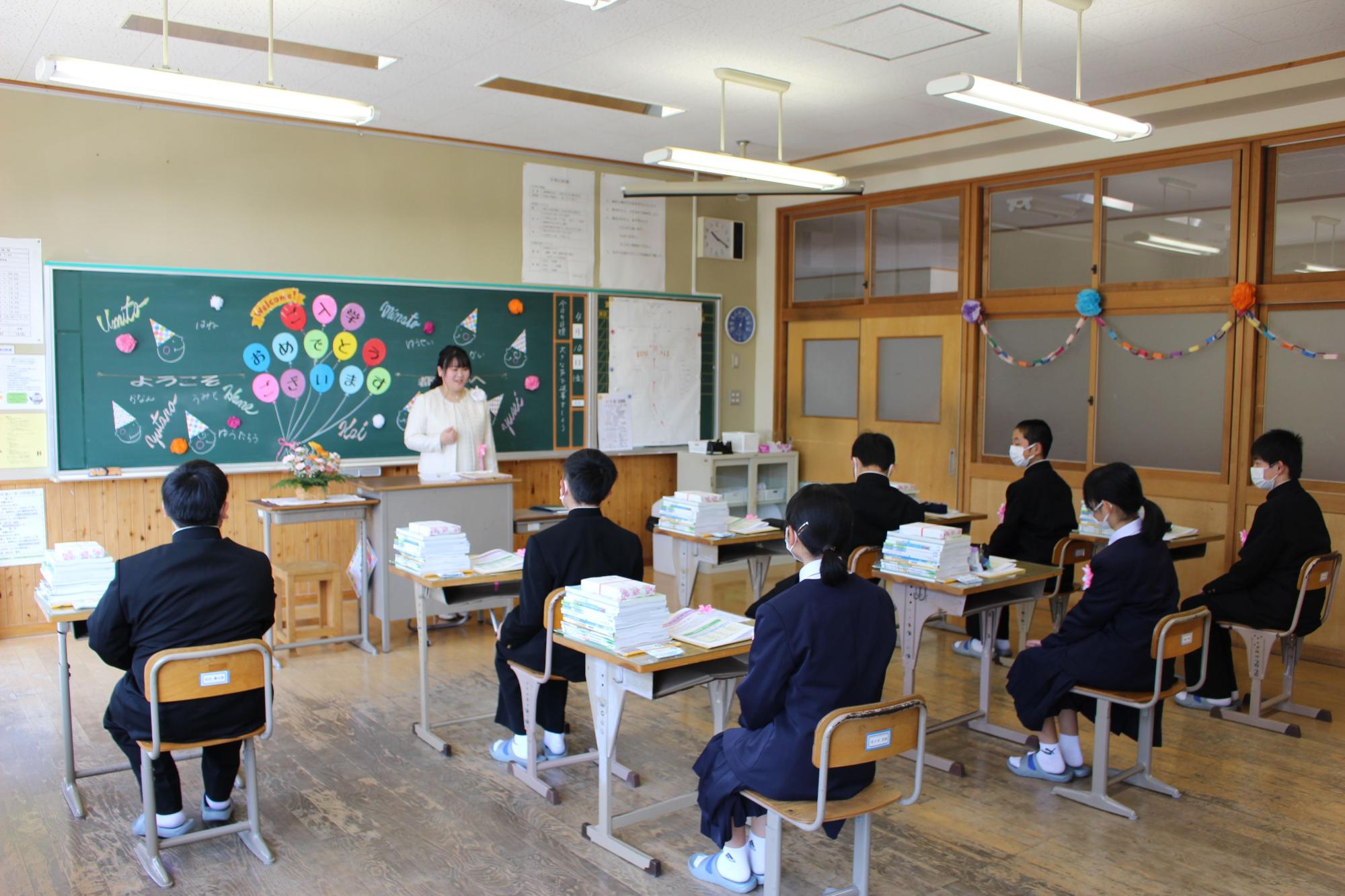 教室で新中学生の男女が沢山の教科書が置かれた机に座り、先生の話を聞いている様子の写真