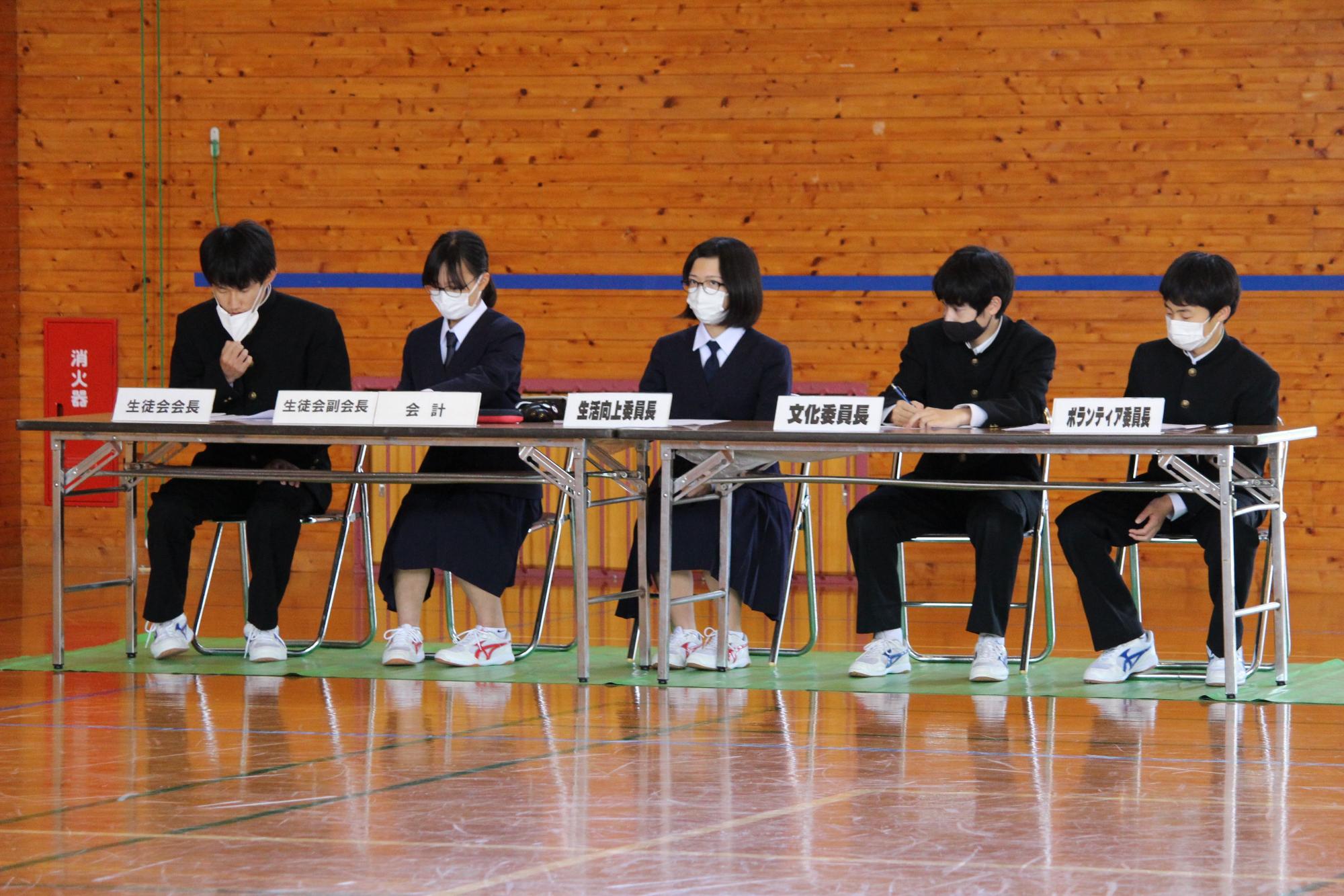 体育館で長机に男子生徒3人と女子生徒2人が並んで座っている写真