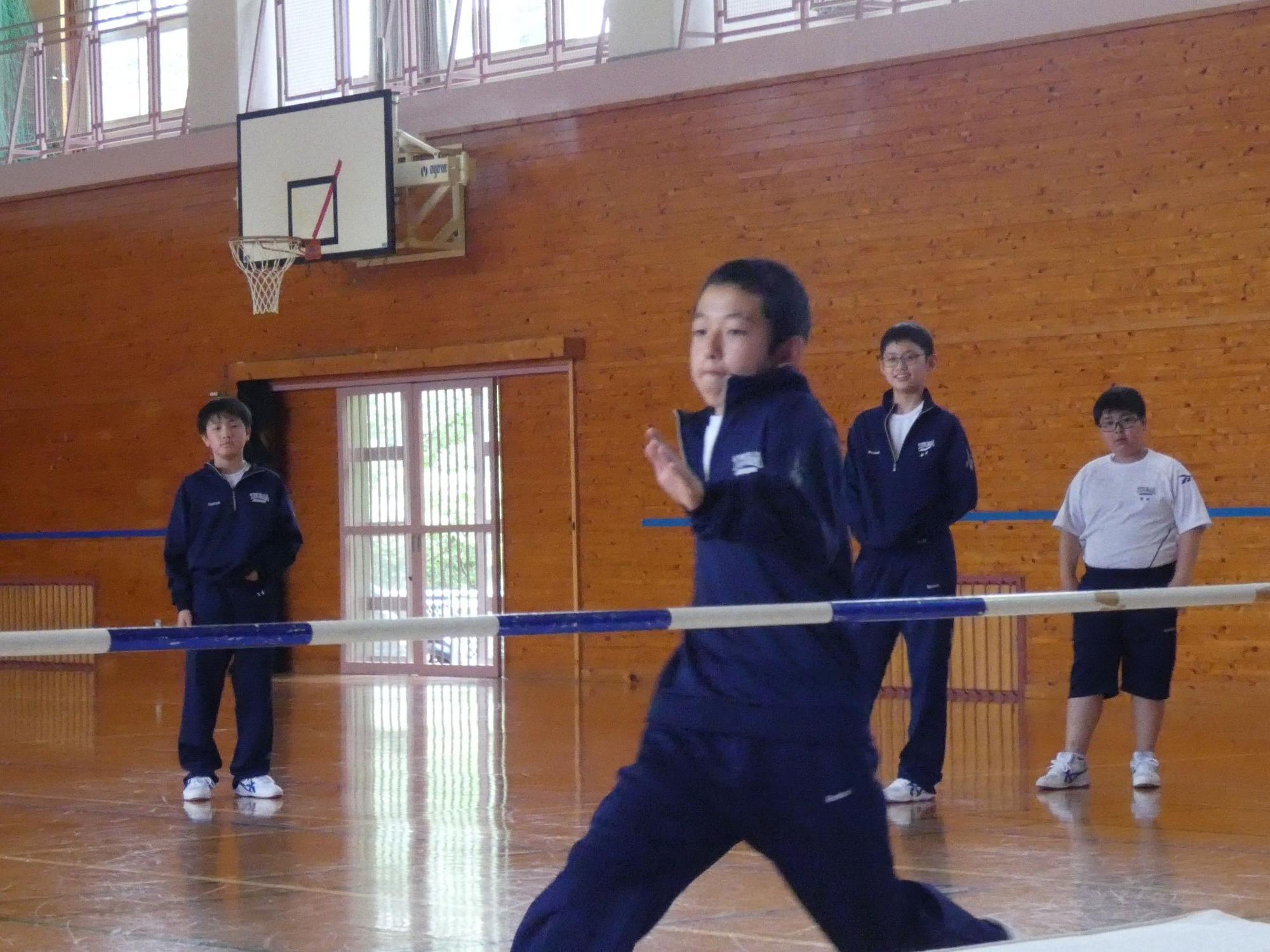 体育館で白と青のポールをジャージ姿の男子生徒が飛び越えようとしている様子の写真