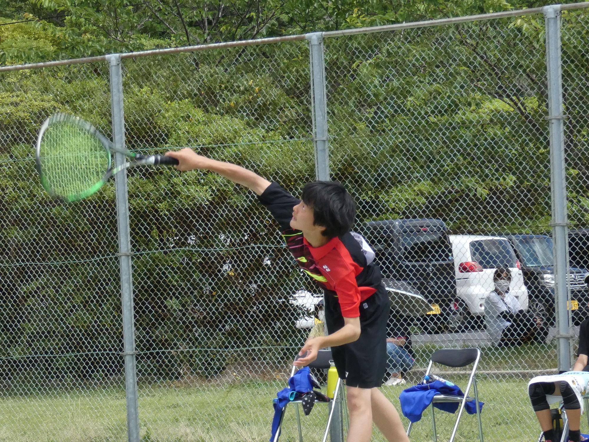 高いフェンスが設置されたテニスコートで、ラケットを振ってサーブを打つユニフォーム姿の男子生徒の写真