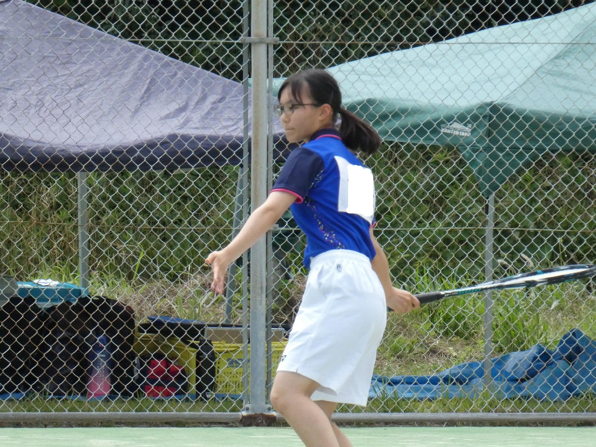 フェンスが設置されたテニスコートでラケットを振りかぶっている眼鏡をかけた女子生徒の写真