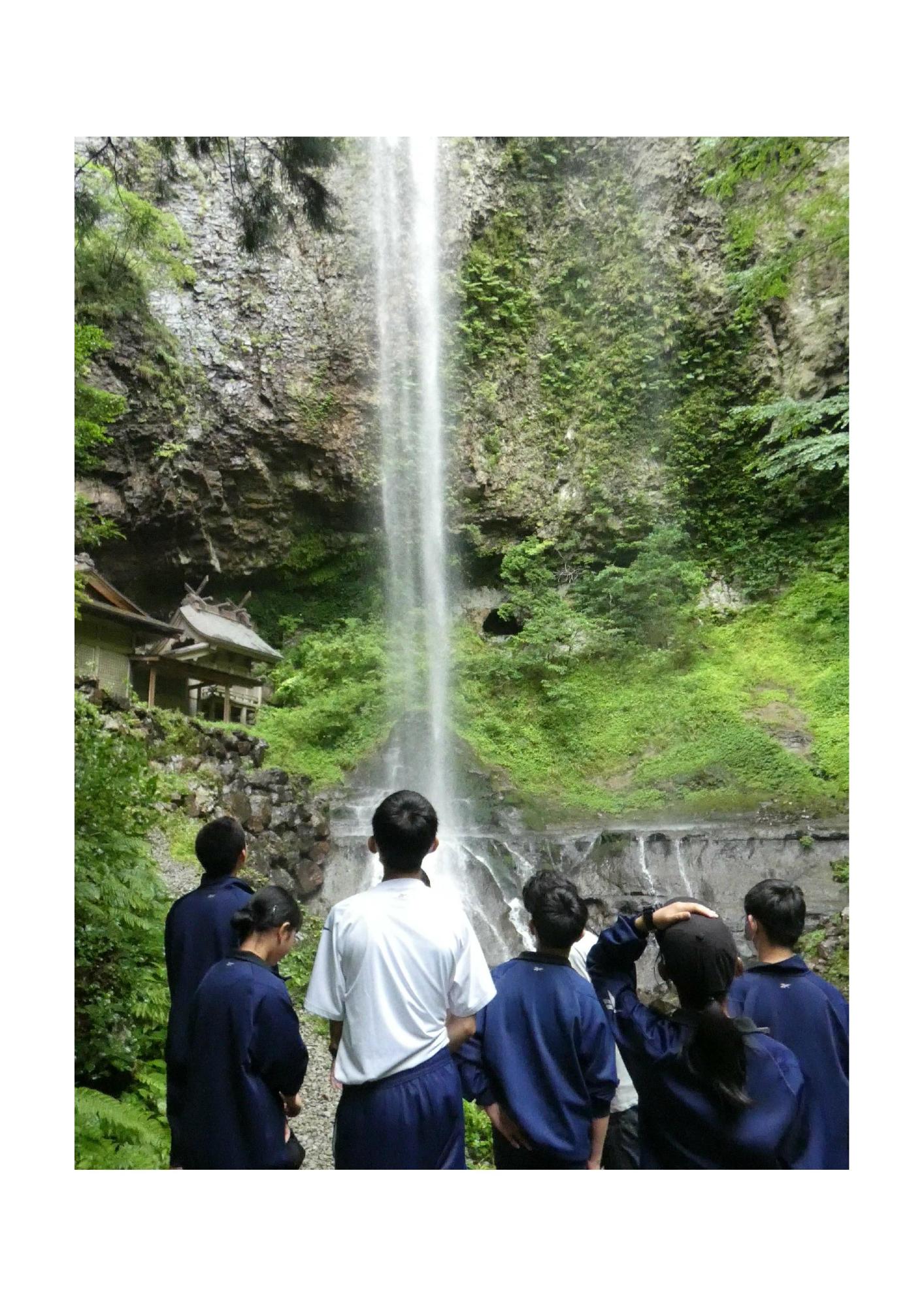 大きな山肌の上から落ちてくる一筋の滝を見上げているジャージ姿の生徒たちの写真