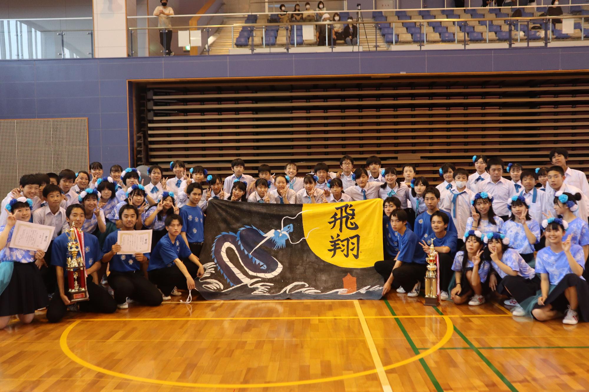 体育館でトロフィーと賞状、横断幕を掲げて記念撮影する生徒たちの写真