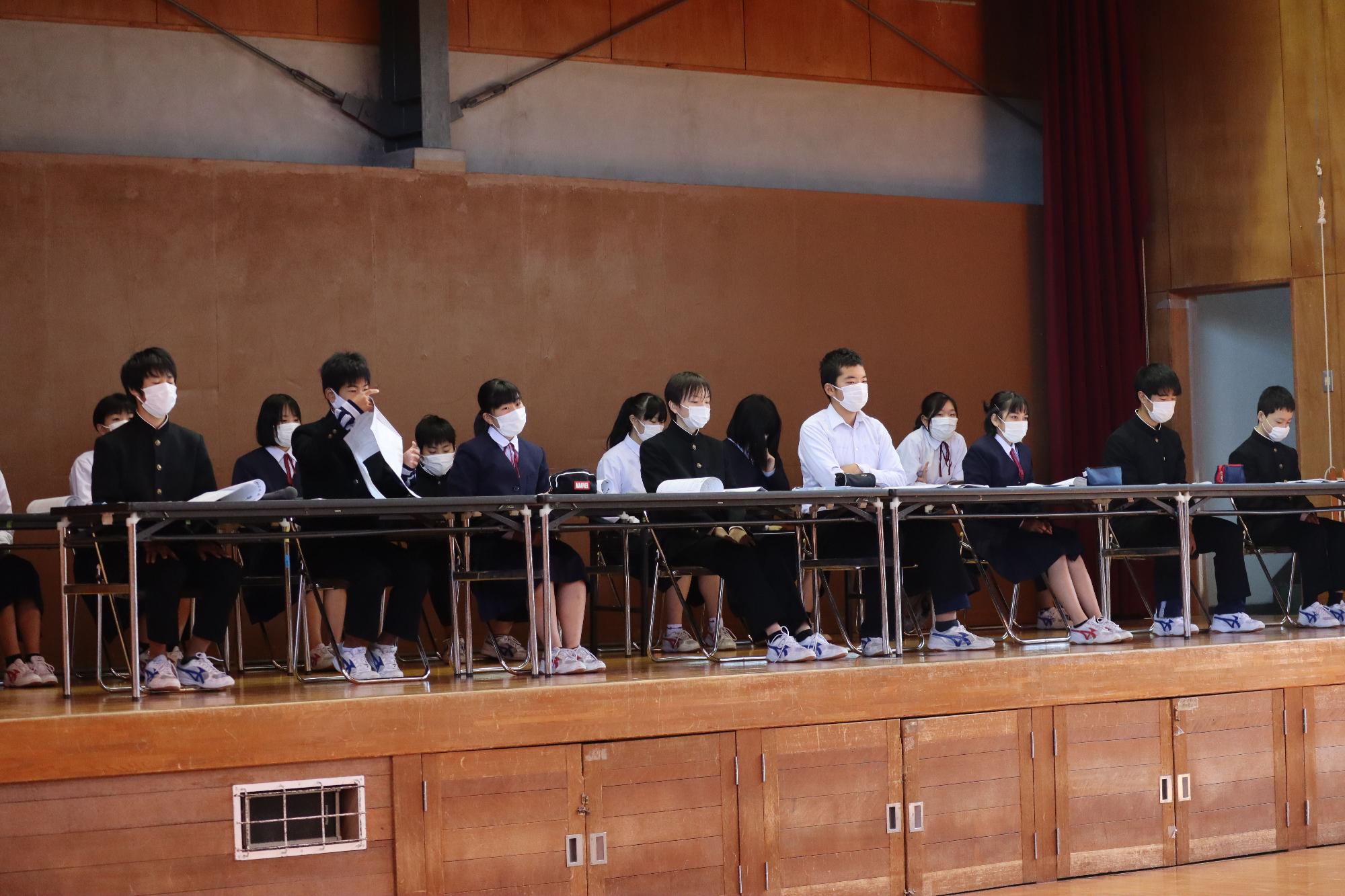 ステージ上に沢山の机が並べられ生徒が何人も座っている写真