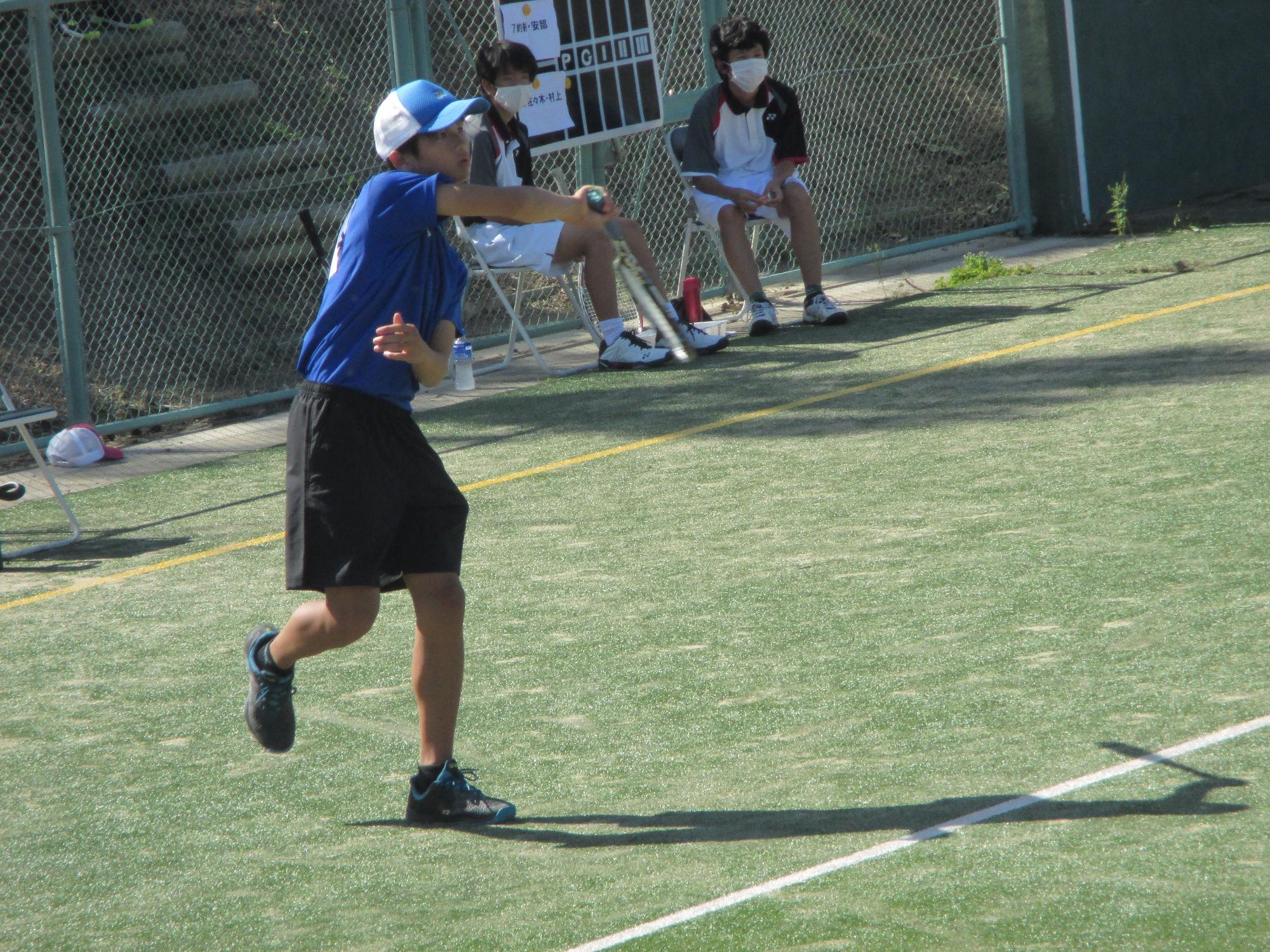 屋外テニスコートで全力でサーブを打つ帽子を被った生徒の写真