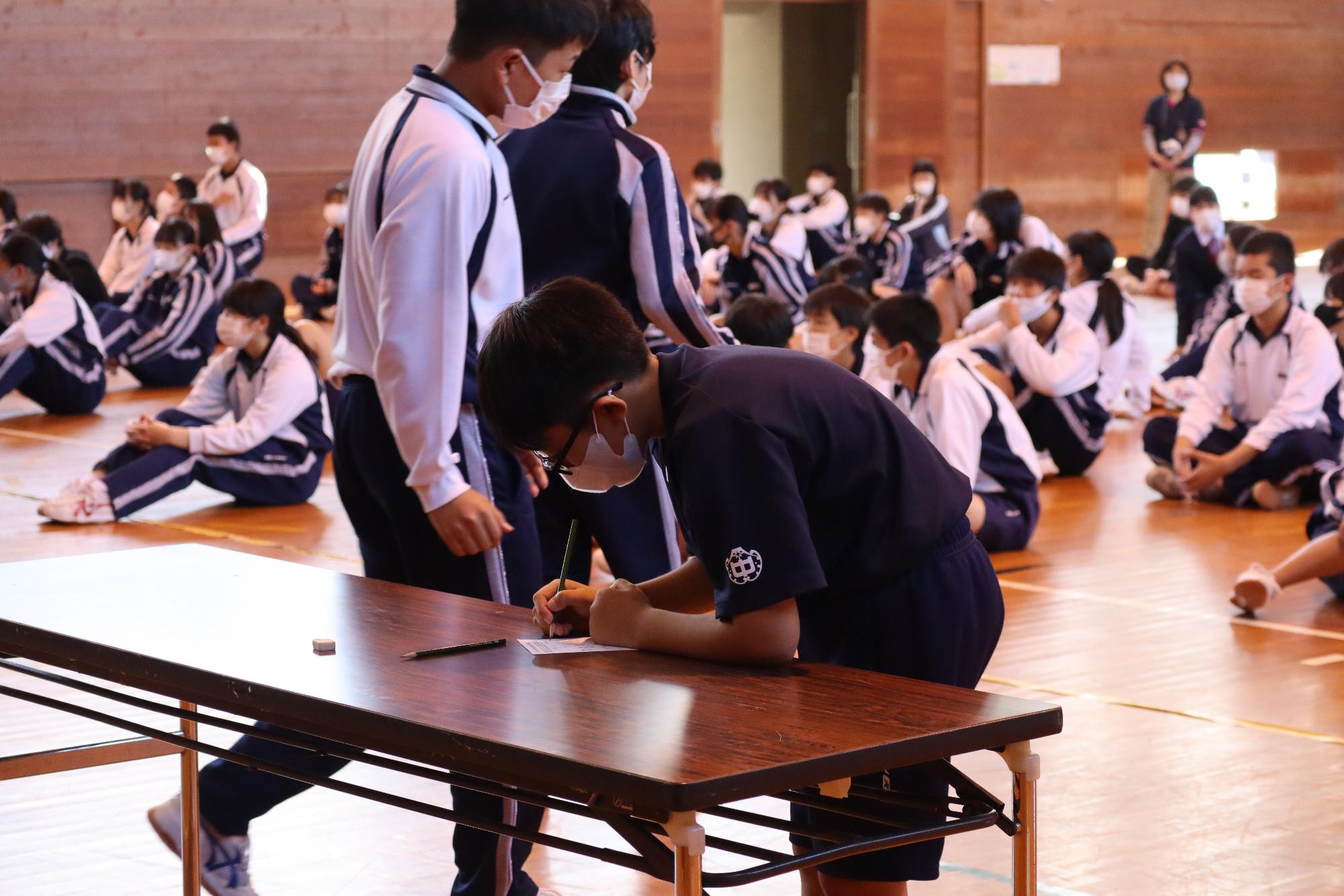 体育館に設置された机で、投票用紙に候補者の名前を記載している生徒の写真