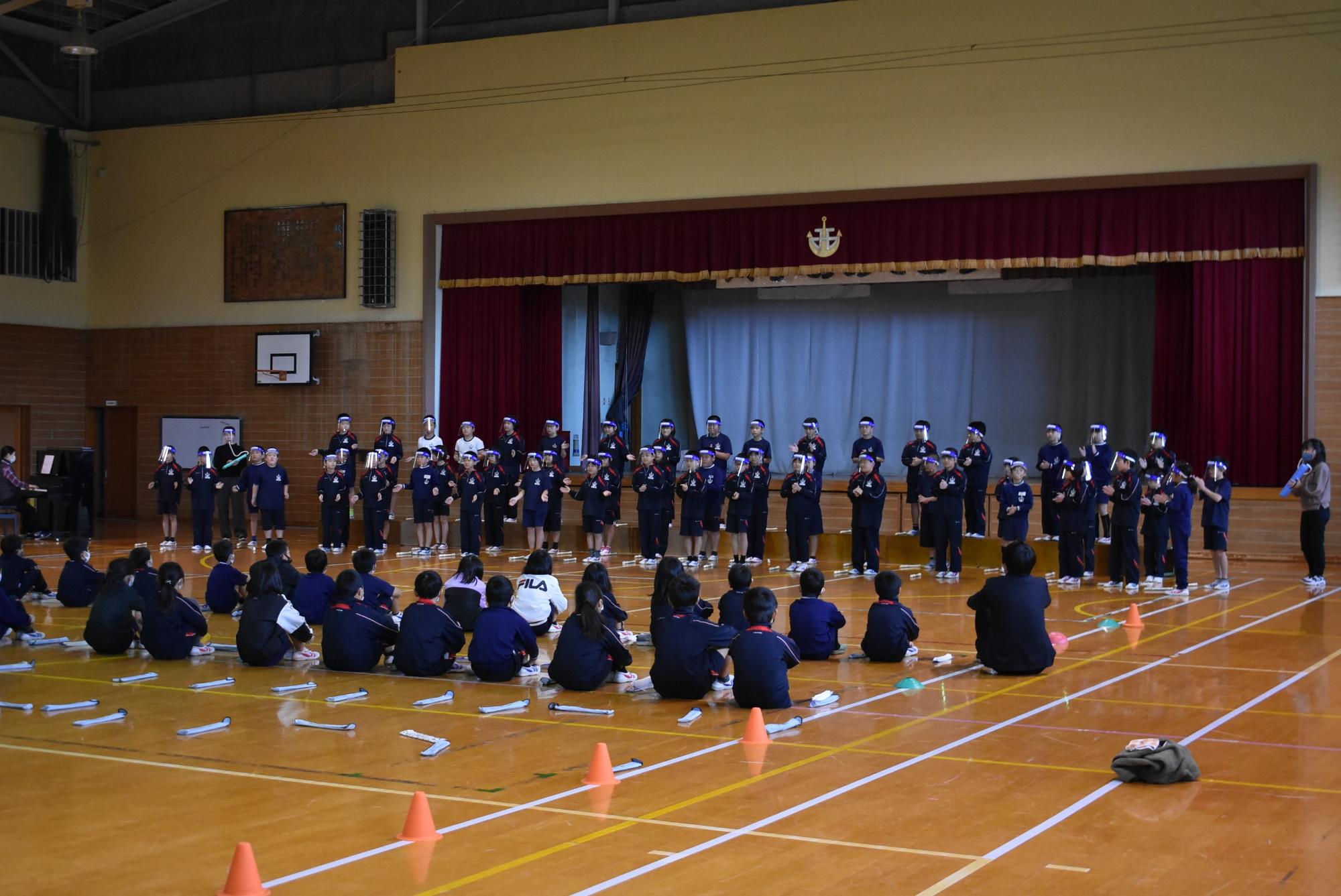 体育館のステージでフェイスシールドを着けて合唱する児童たちの写真