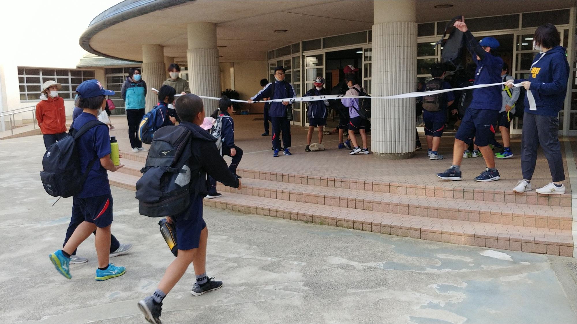 白いゴールテープが貼られた昇降口前で大きな荷物を背負った生徒たちが歩いてくる写真