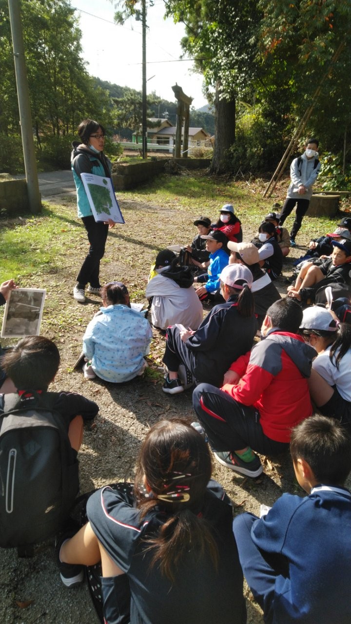 道路の脇のスペースに座り、地図を持って説明する先生の話を座って聞いている生徒たちの写真