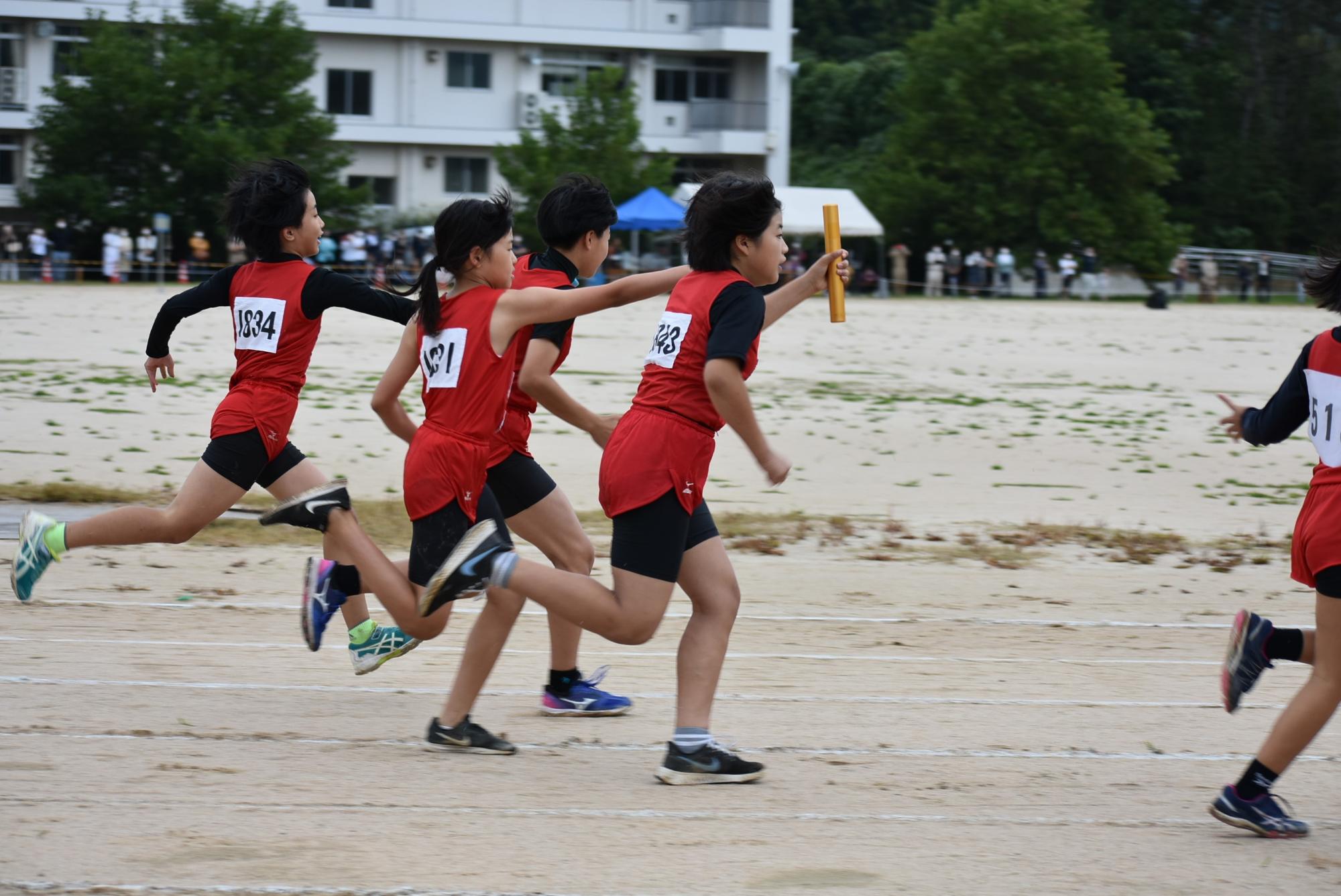グラウンド上のトラックで赤色のユニフォームを着用した生徒がリレーのバトンを全力でつなぐ写真