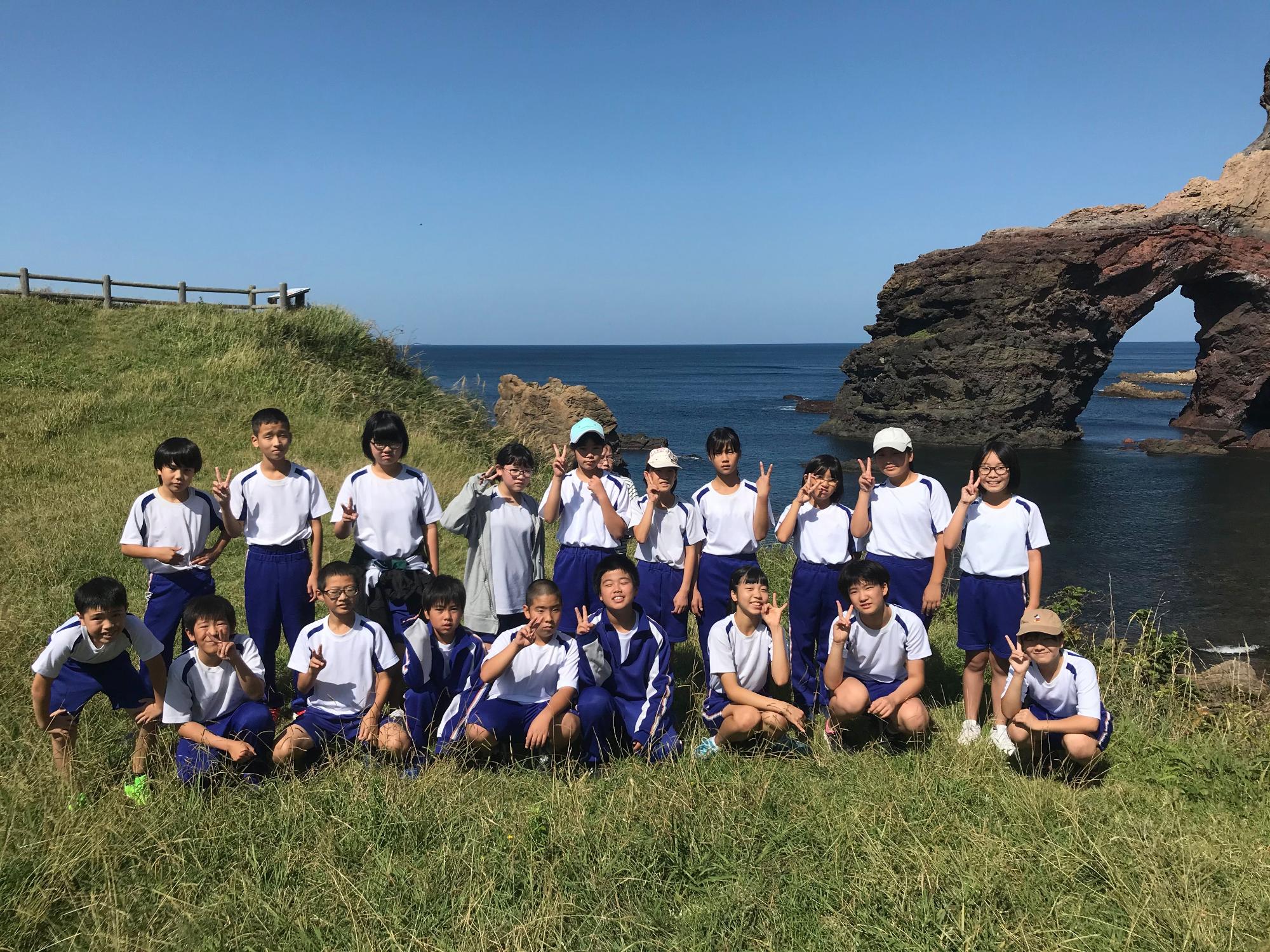 奥に特徴的な岸壁がある岬で2列になって写真に映る児童たちの写真