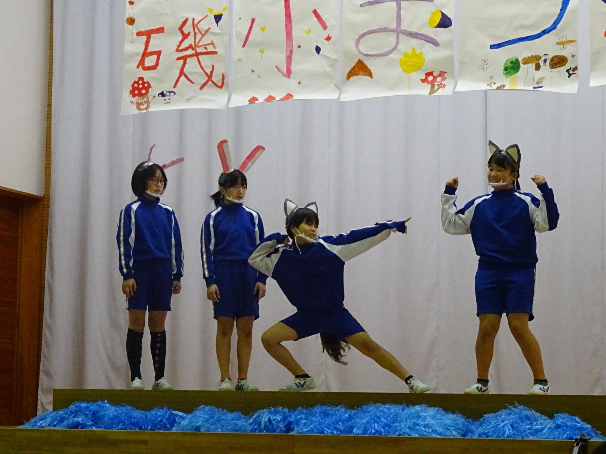大小様々な耳の飾りをつけた児童4人がステージ上に立ち、うち一人がポーズを取っている様子の写真