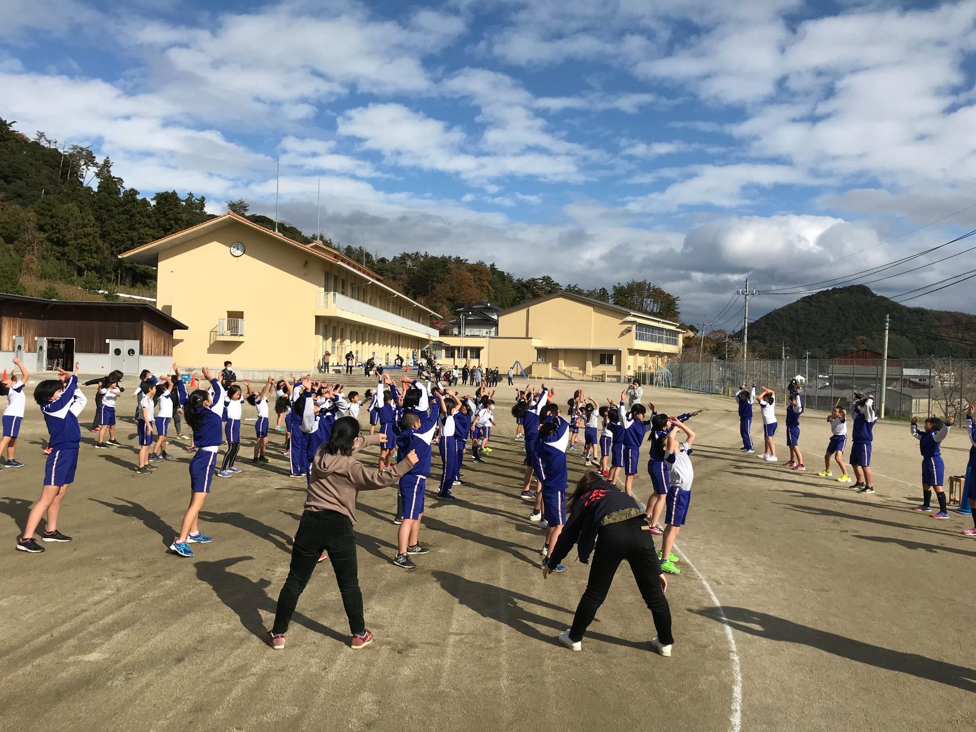 雲がまばらにある青空の下、体操着姿の児童たちが体を大きく回して準備体操をしている様子の写真