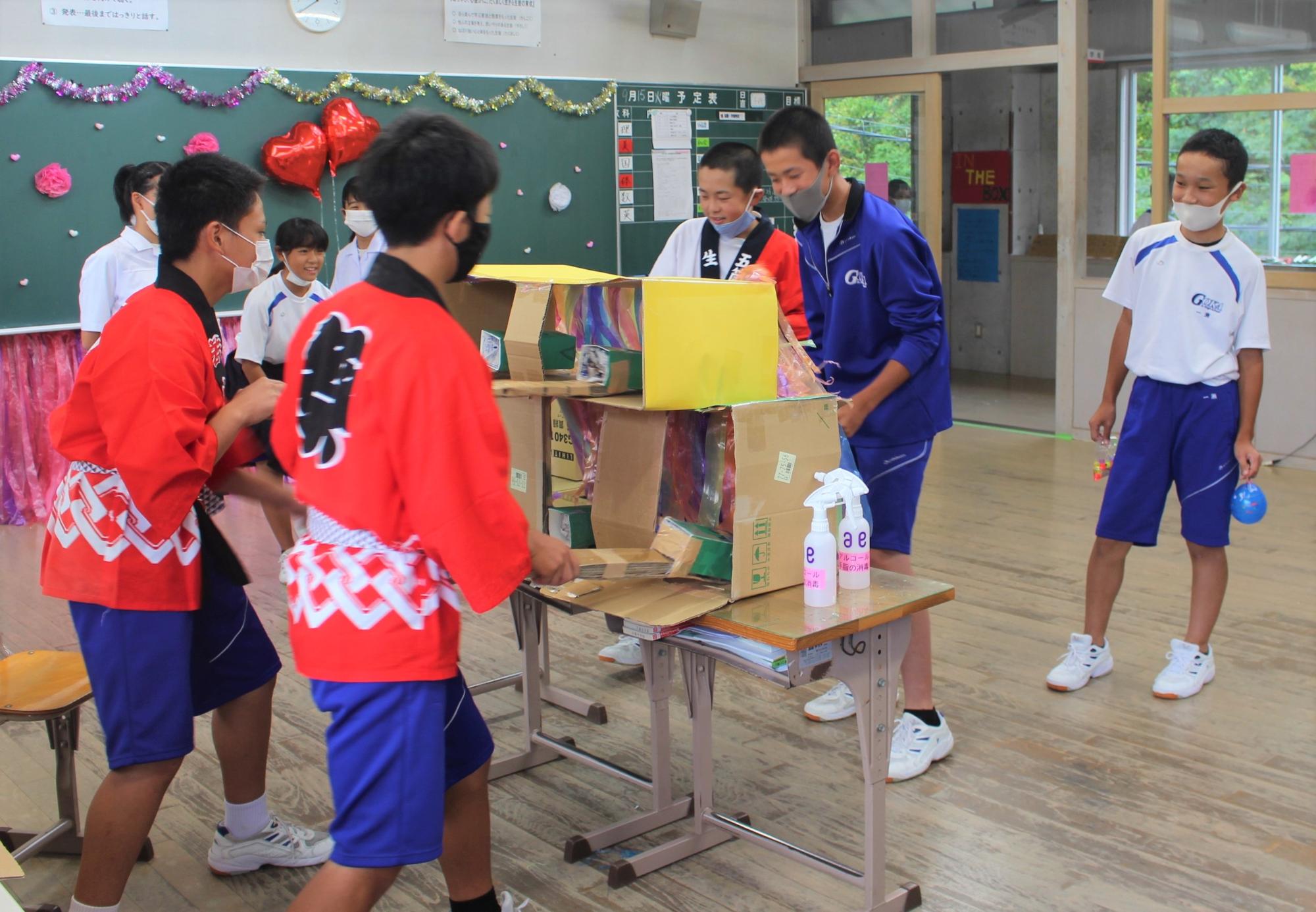 赤色の法被を着た児童や体育着を着た生徒が教室内で手造りの屋台ゲームで遊んでいる様子の写真