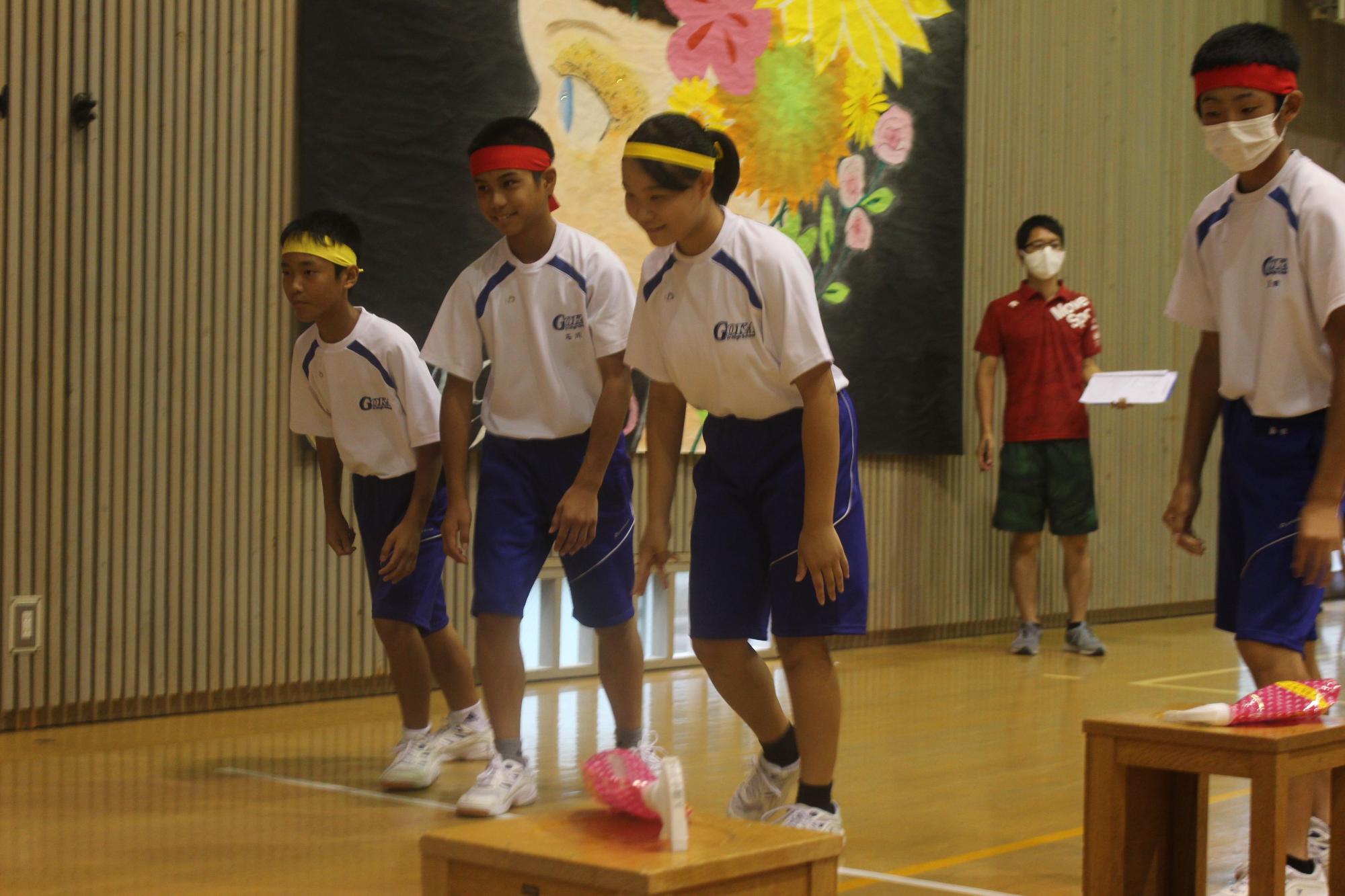 黄色や赤色のハチマキを頭に巻いた体操着姿の複数人の生徒たちが横に並んで立っている様子の写真